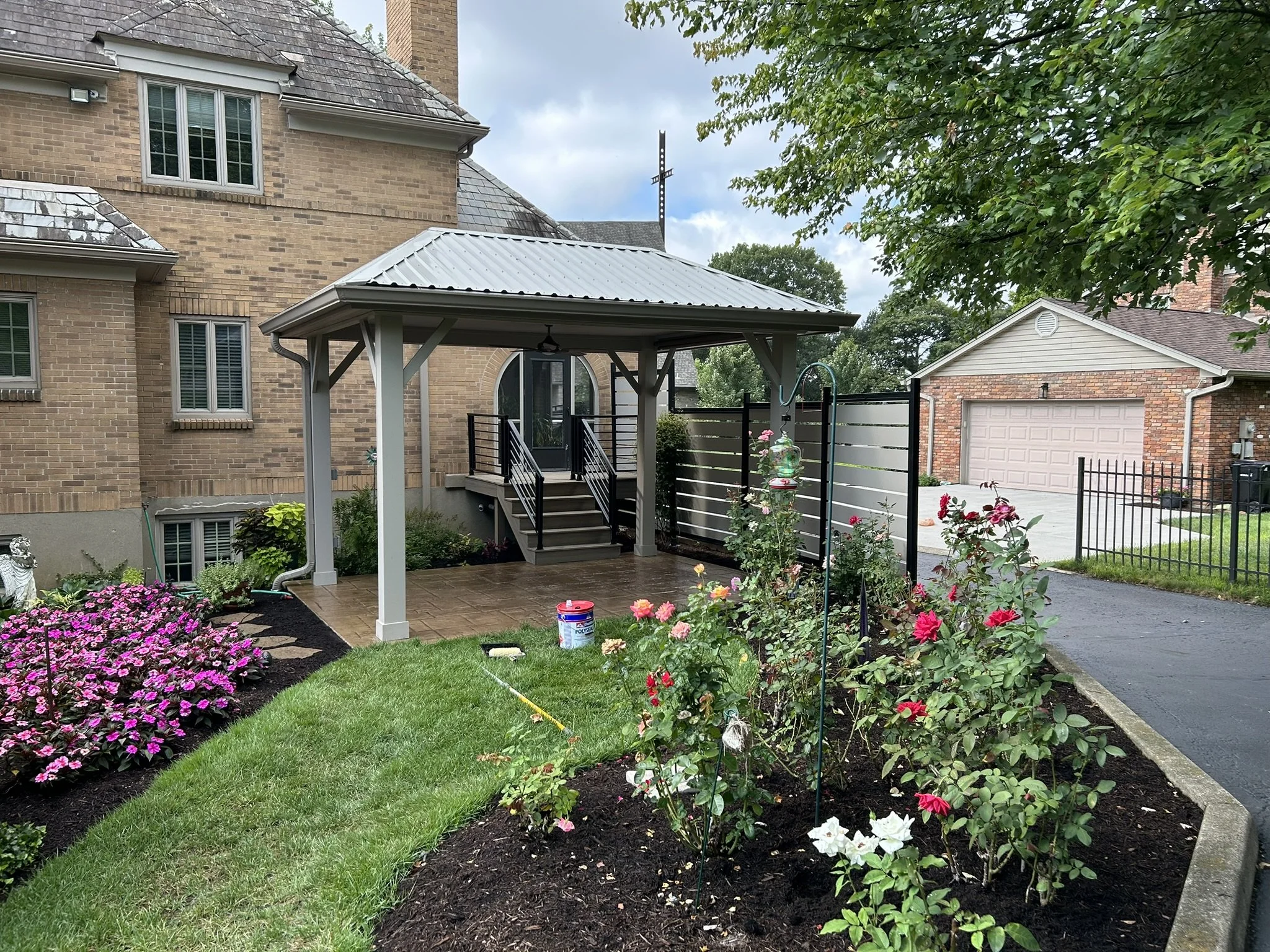 A backyard garden with blooming pink and white flowers along a green lawn, a small flower bed with purple flowers, a covered patio with stairs, a house with brick walls and multiple windows, a gray gate, and a garage in the background under cloudy sk