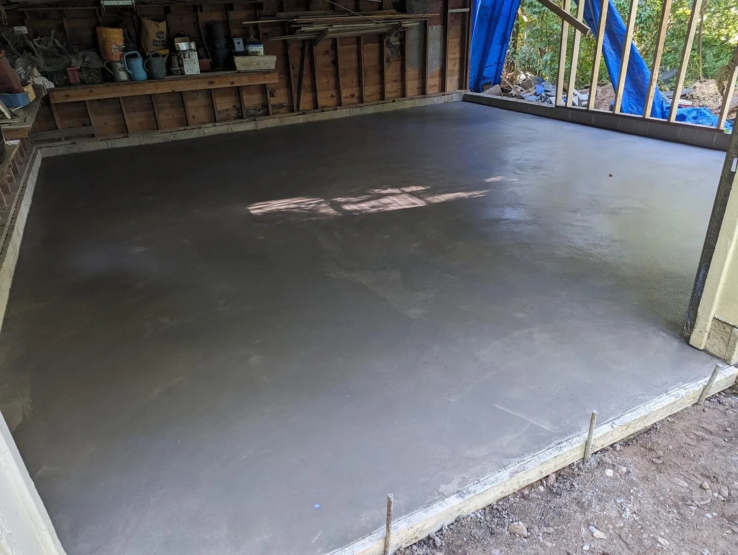 Freshly poured concrete slab in a construction garage or workshop, with tools and materials stored on walls, and some exterior construction visible. The concrete appears smooth and wet.