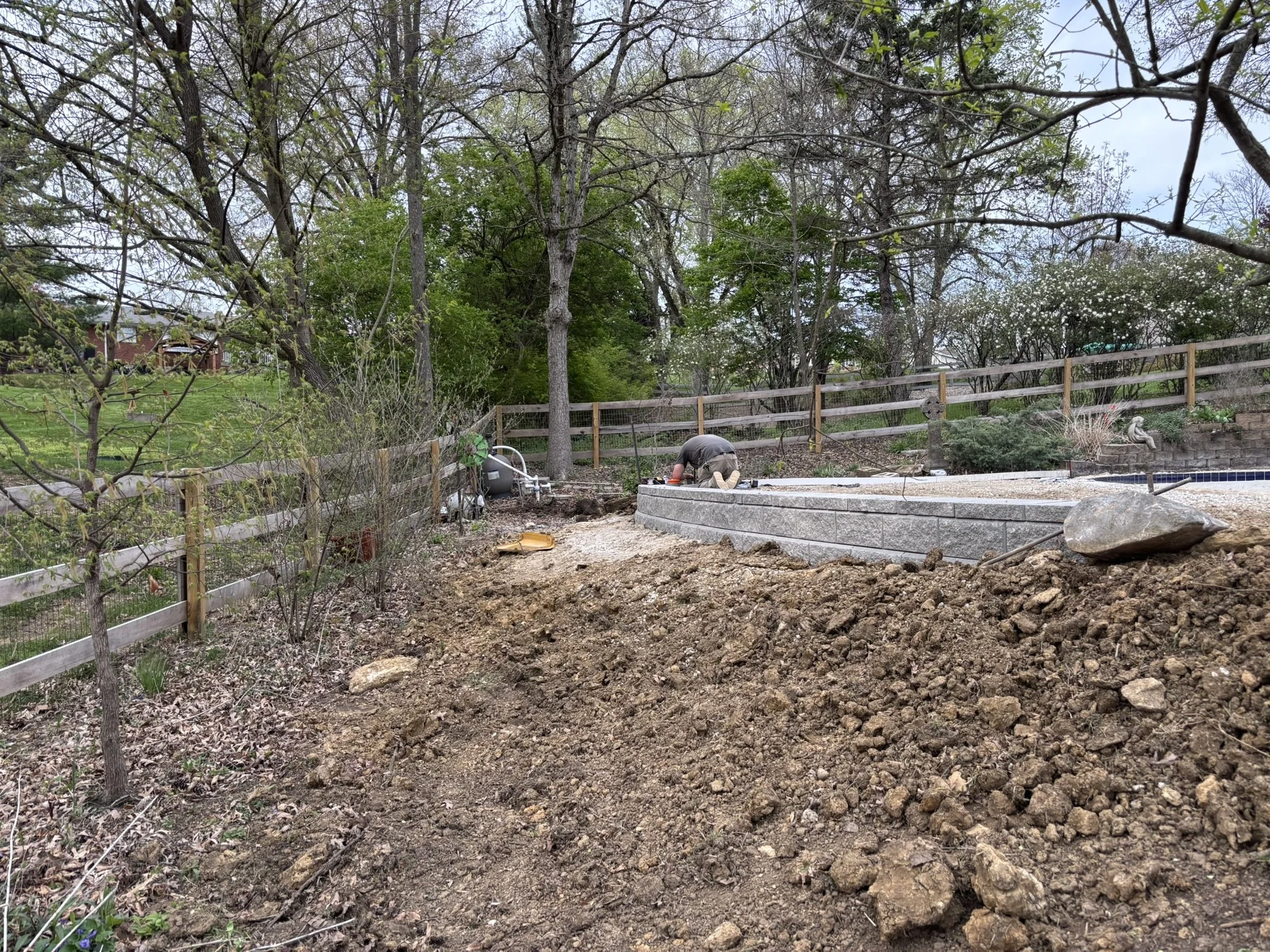 A backyard with trees, a wooden fence, and a man working on a construction project near a kidney-shaped pool.
