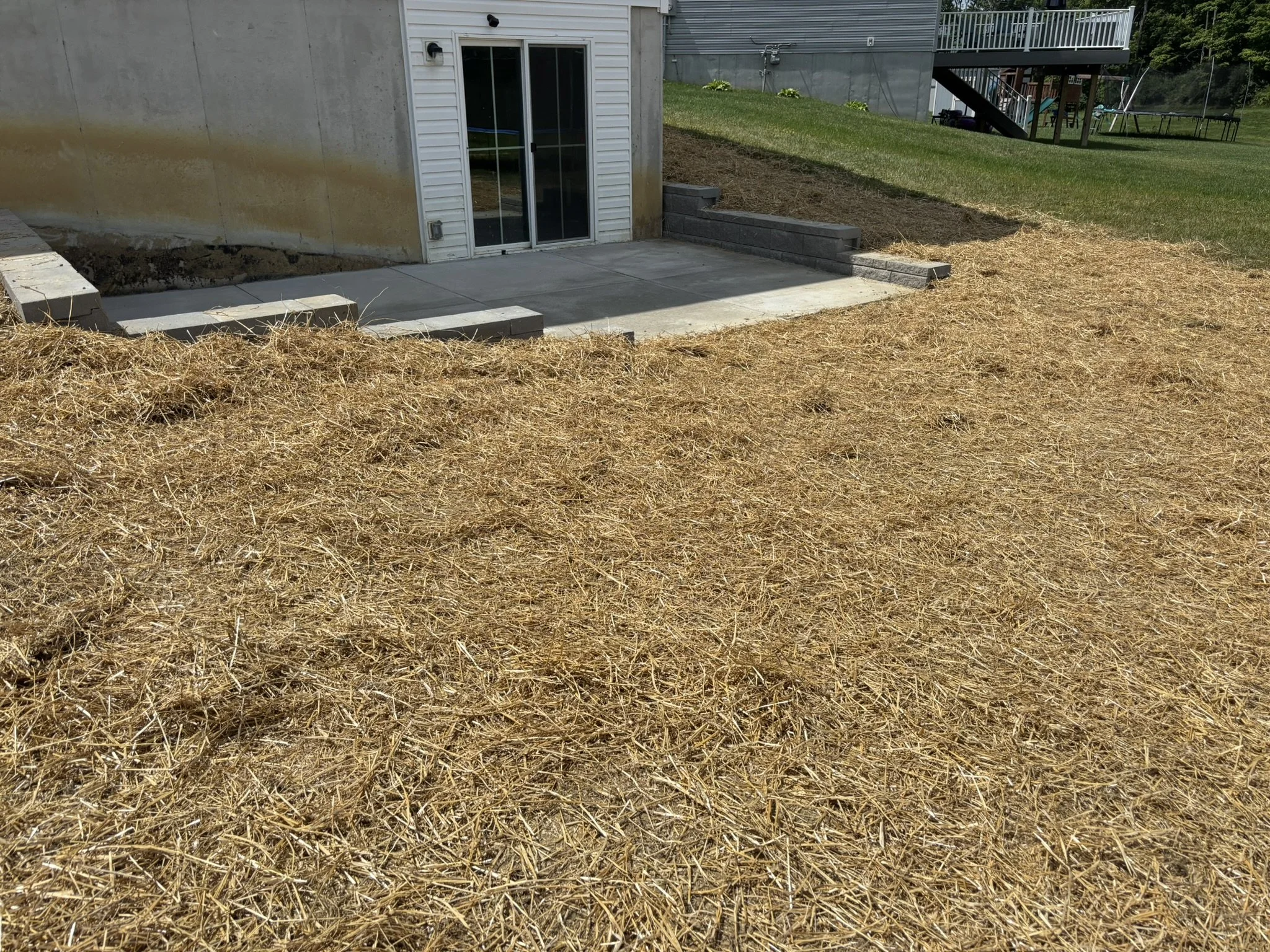 A backyard patio area with a concrete slab leading to a sliding glass door. The door is part of a house with white vinyl siding and a concrete foundation. There are small steps made of concrete blocks and pavers leading up to a grassy lawn. The yard 