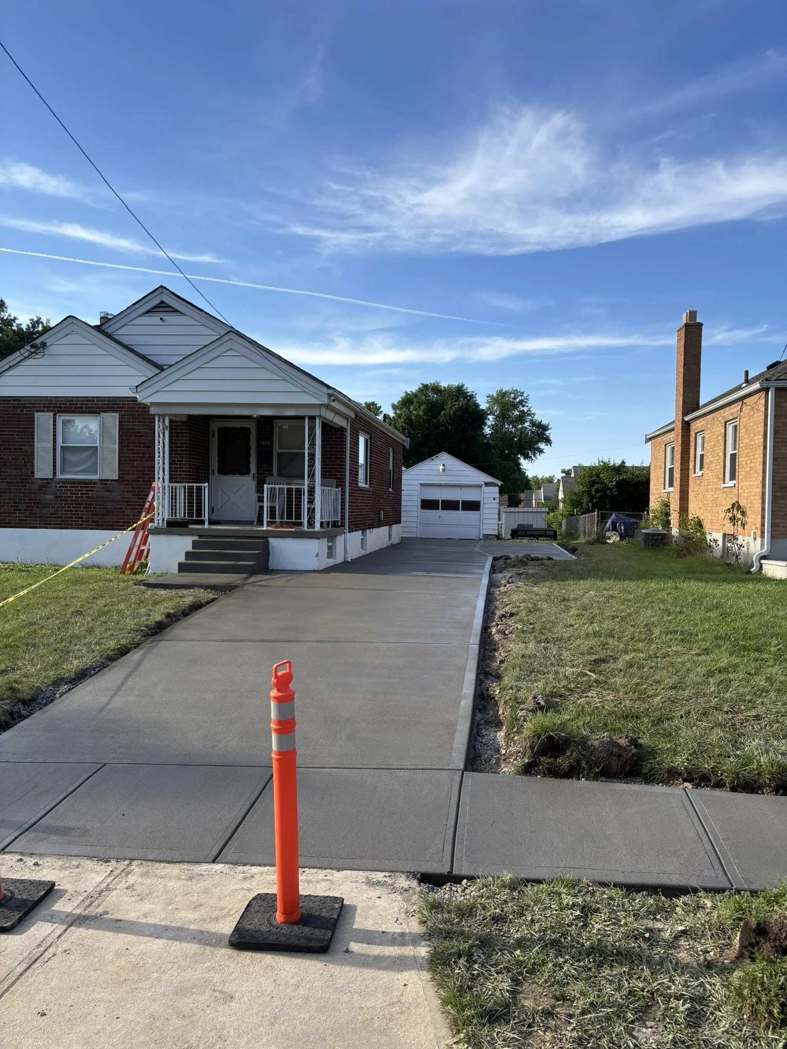 A newly poured concrete sidewalk leading to the front porch of a house, with an orange safety cone and caution tape in the foreground, and a house with white siding and brick on the left side, and another brick house on the right side under a blue sk