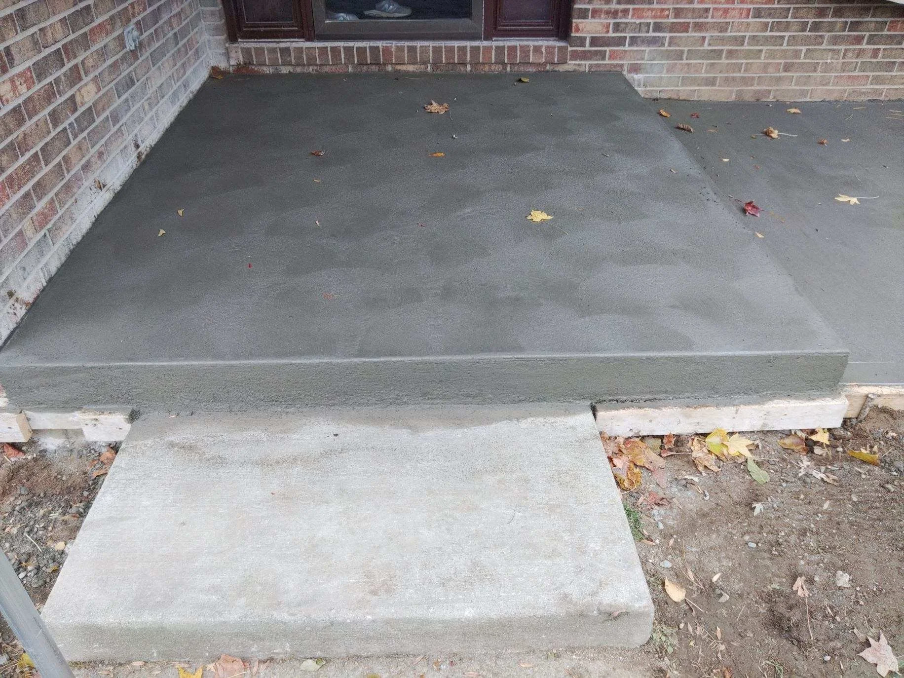 Concrete porch with two steps in front of a brick house, with fallen leaves scattered on the surface.