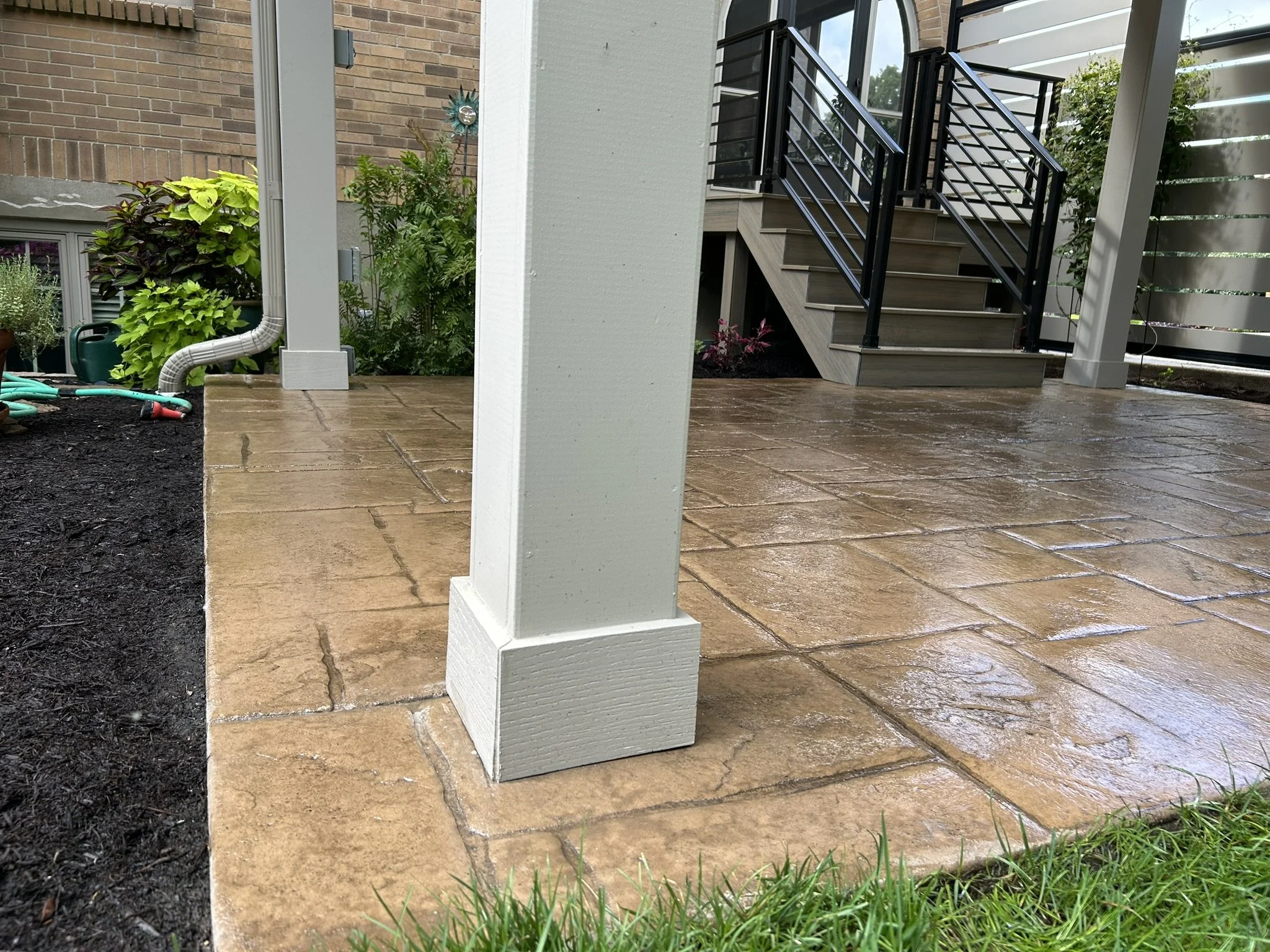 Close-up of a backyard patio with wet stone tiles, a white wooden pillar, garden plants, and metal stairs with black railings leading to an upstairs porch.
