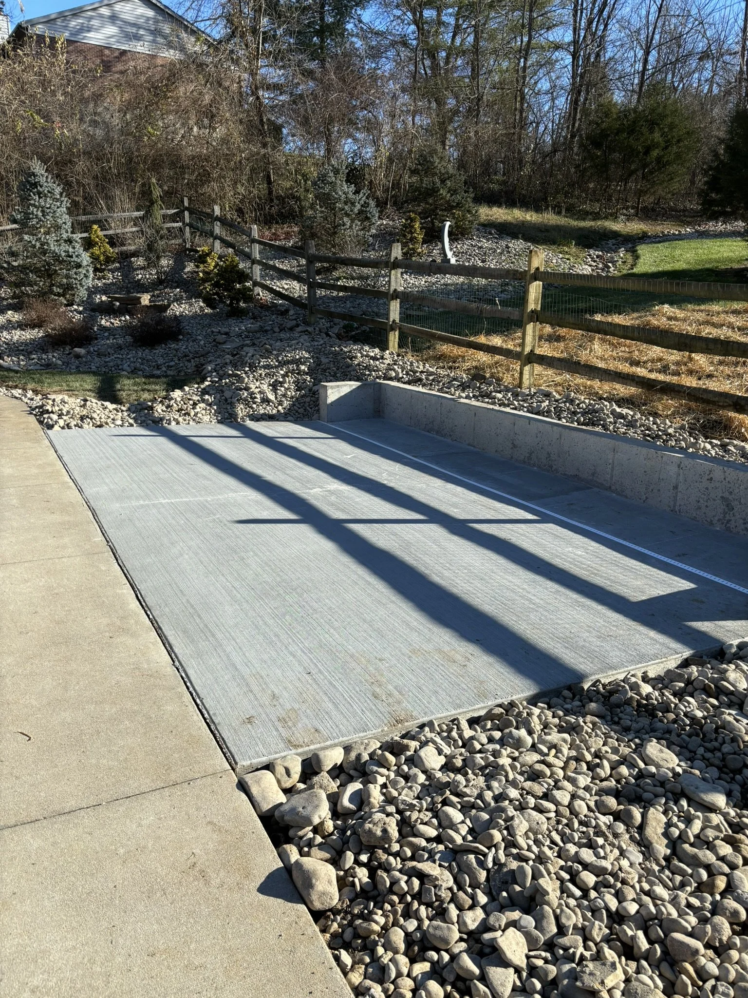 A garden with various small trees and bushes, a wooden fence, and a concrete patio with a rock garden, under a blue sky with a house partially visible in the background.