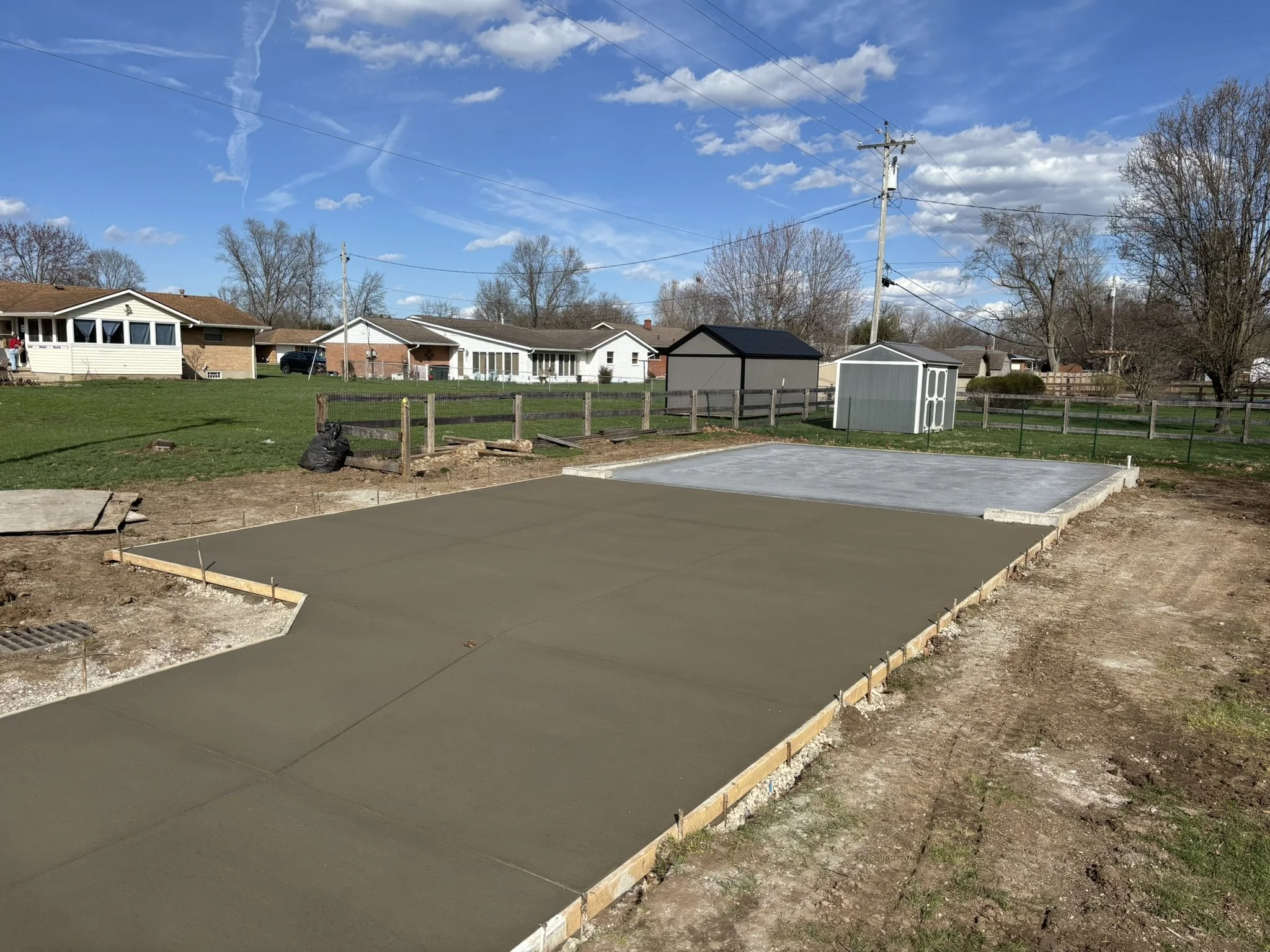 A backyard with a concrete patio area under construction, surrounded by a wood frame, in front of a fenced backyard with grass, garden beds, small garden shed, and neighboring single-story houses under a blue sky with clouds and power lines.