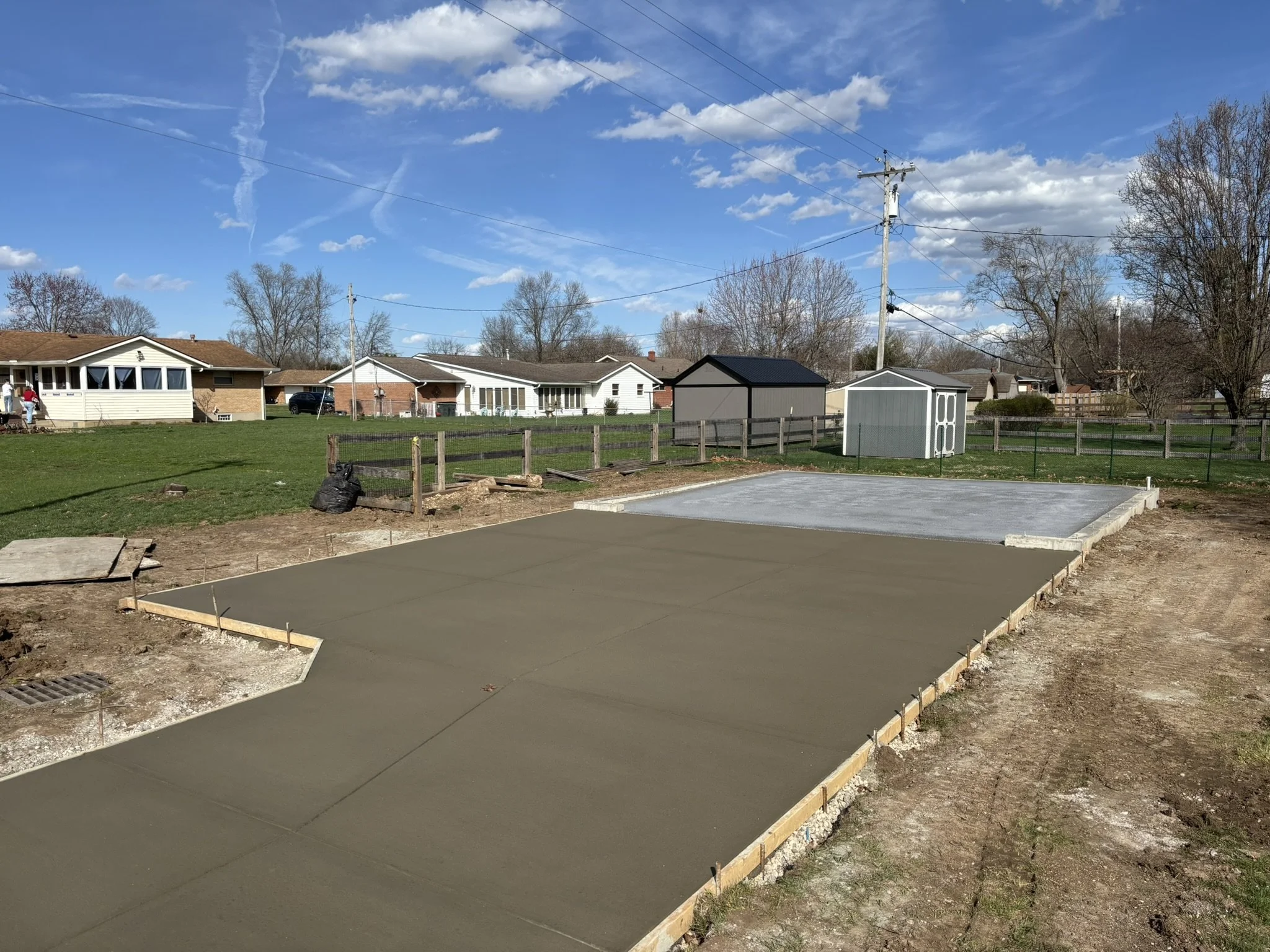 A backyard with a concrete slab foundation and wooden framing for a patio or sidewalk, with a grassy yard and houses in the background under a clear blue sky with clouds and powerlines.