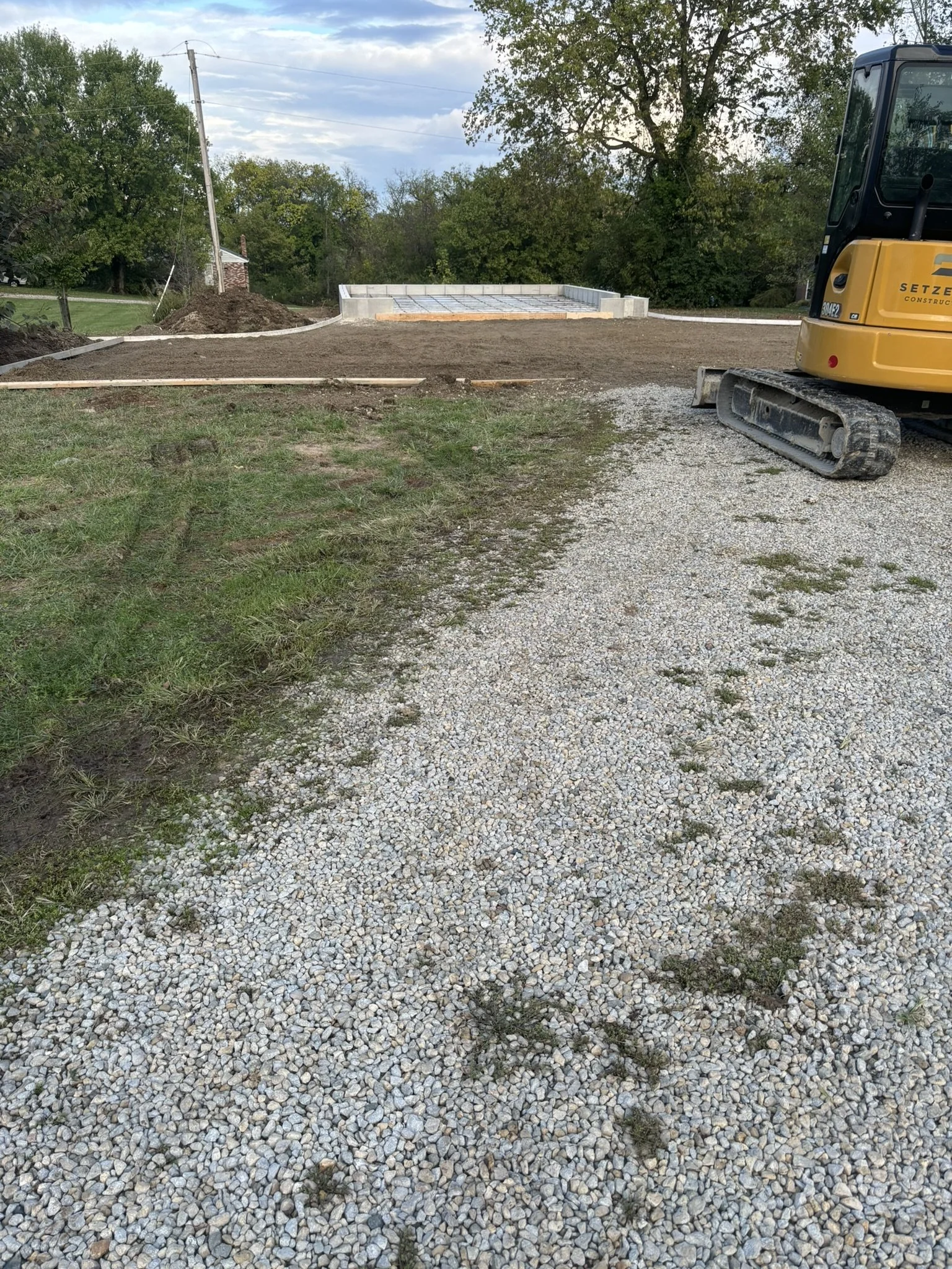A construction site with a small yellow excavator on the right and a concrete foundation with a rebar grid in the center. The ground is a mix of grass, dirt, and gravel, with a wooden frame outlining the construction area. Trees and a cloudy sky are 