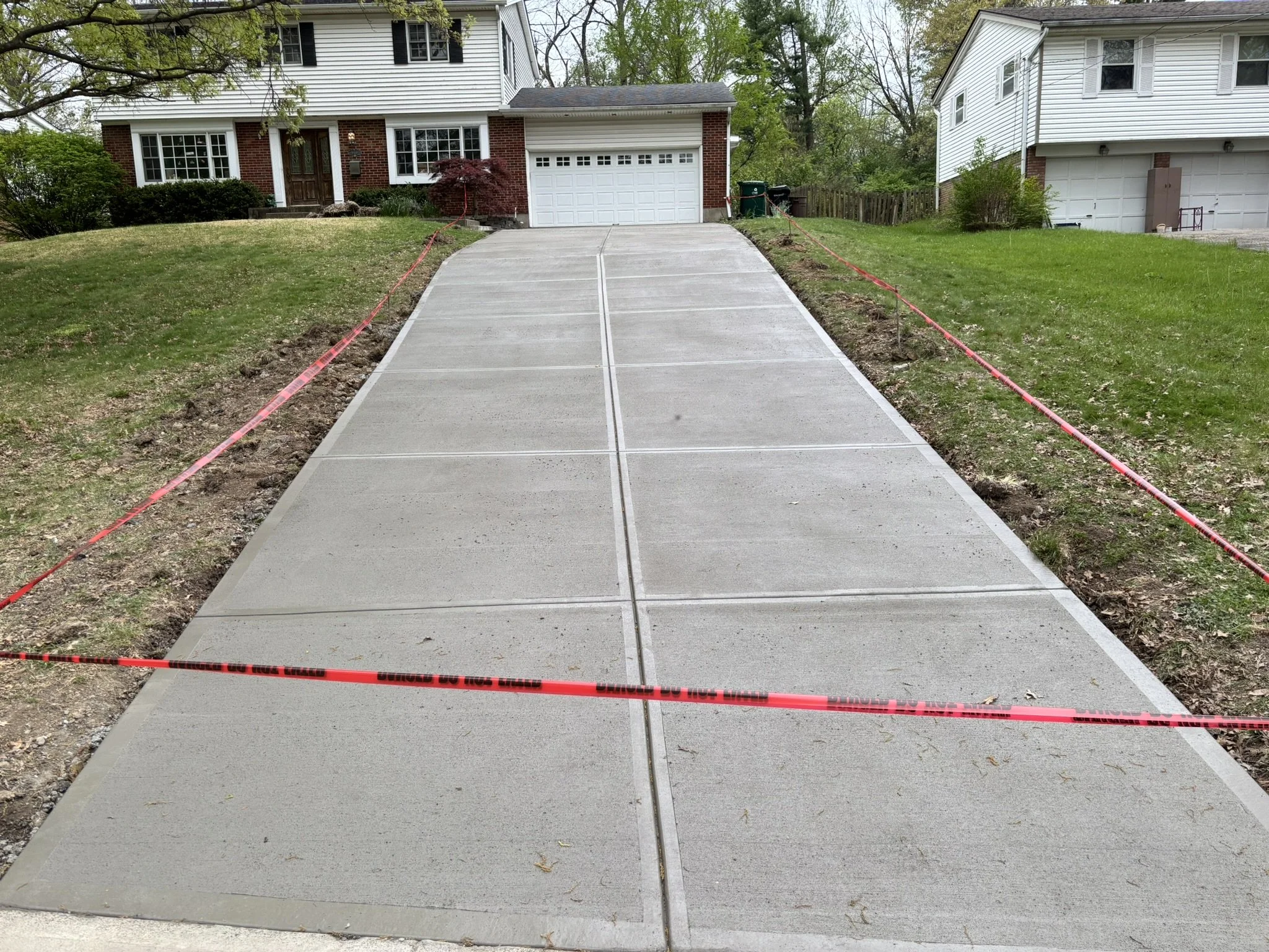 Newly poured concrete driveway in front of a house with two stories, brick and white siding, parked at a slight incline, with orange caution tape around the edges indicating construction.