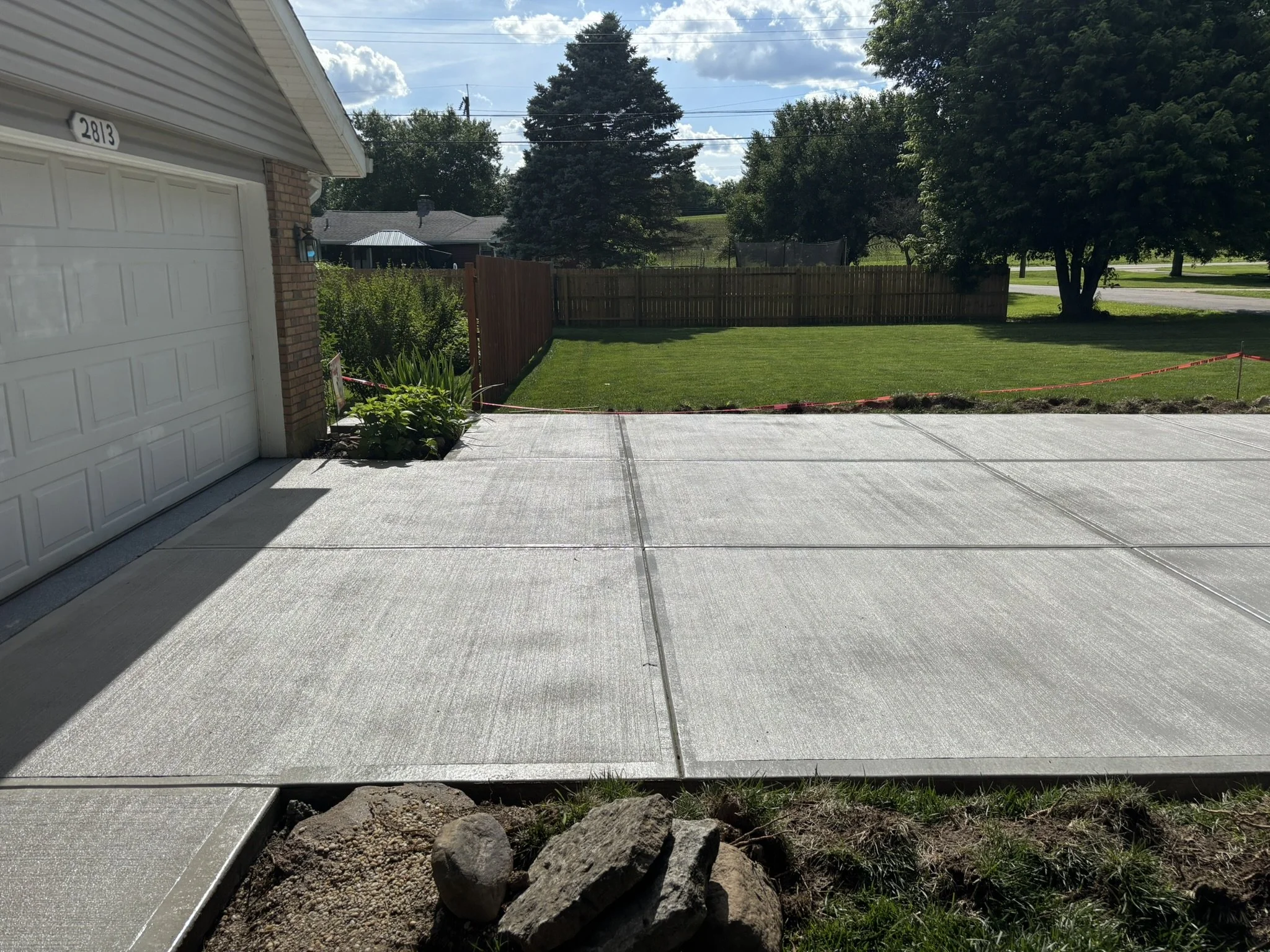 A suburban backyard with a freshly poured concrete patio, a lawn, trees, a wooden fence, and a clear sky with some clouds.