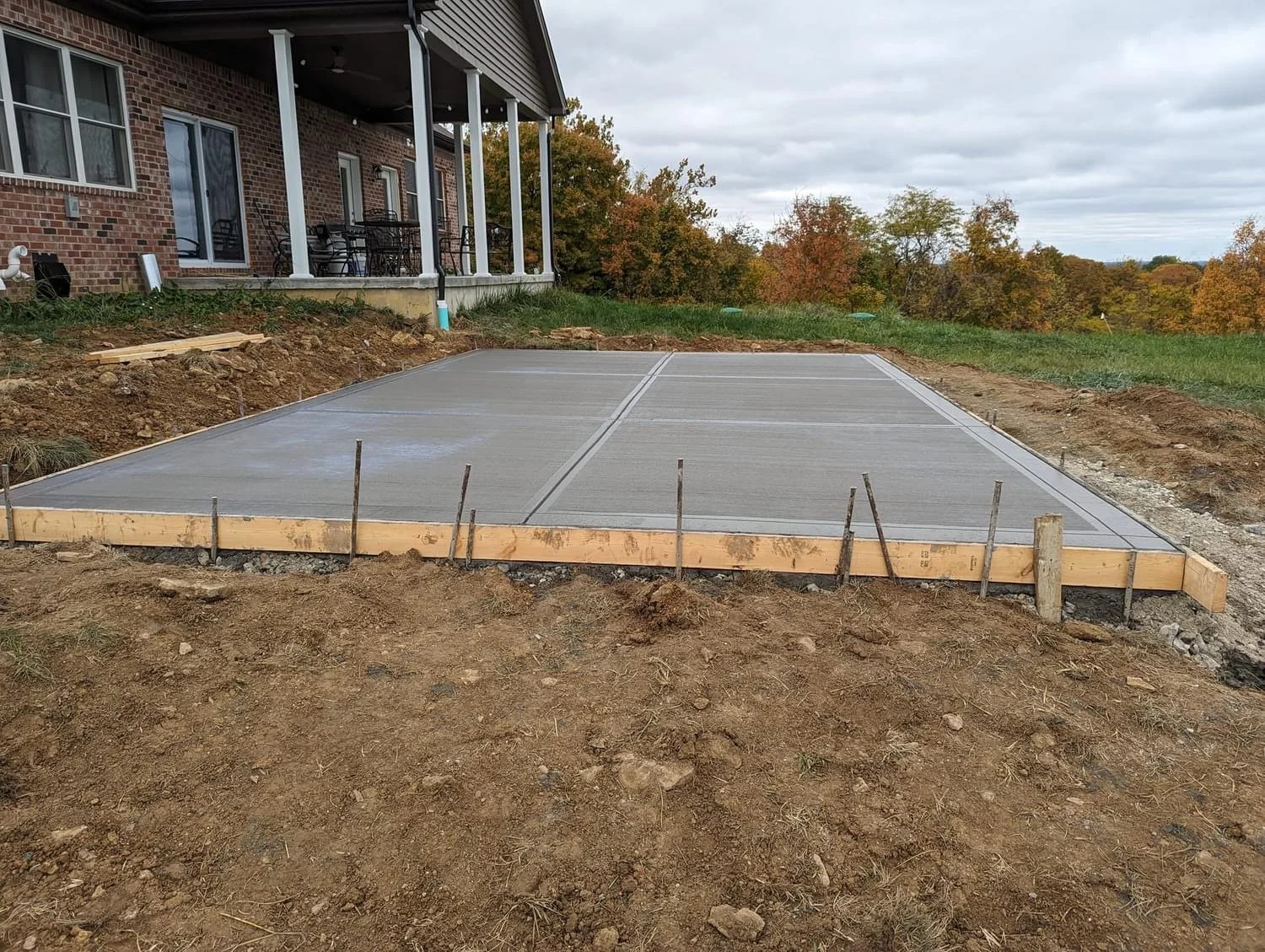 Concrete slab foundation prepared for a new construction site in a backyard, with a brick house and porch on the left, and trees with fall foliage in the background.