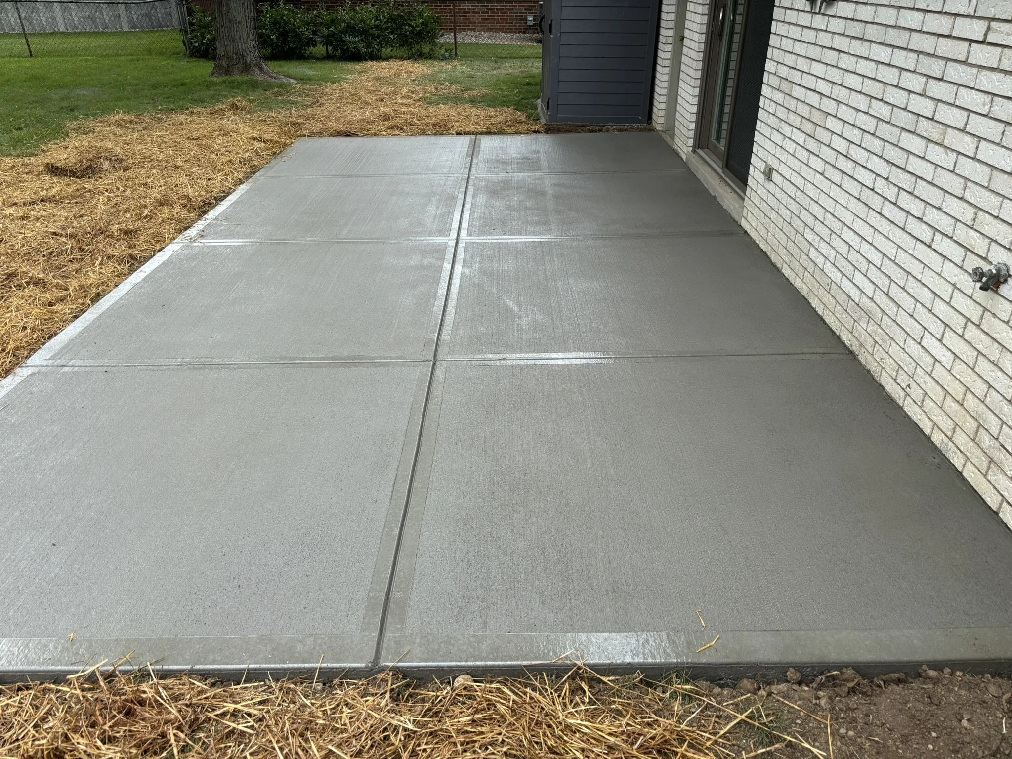 Concrete porch with a matching concrete sidewalk, part of a house with brick walls, a black shutter, and outdoor water spigots; brown mulch and grass with a tree in the background.