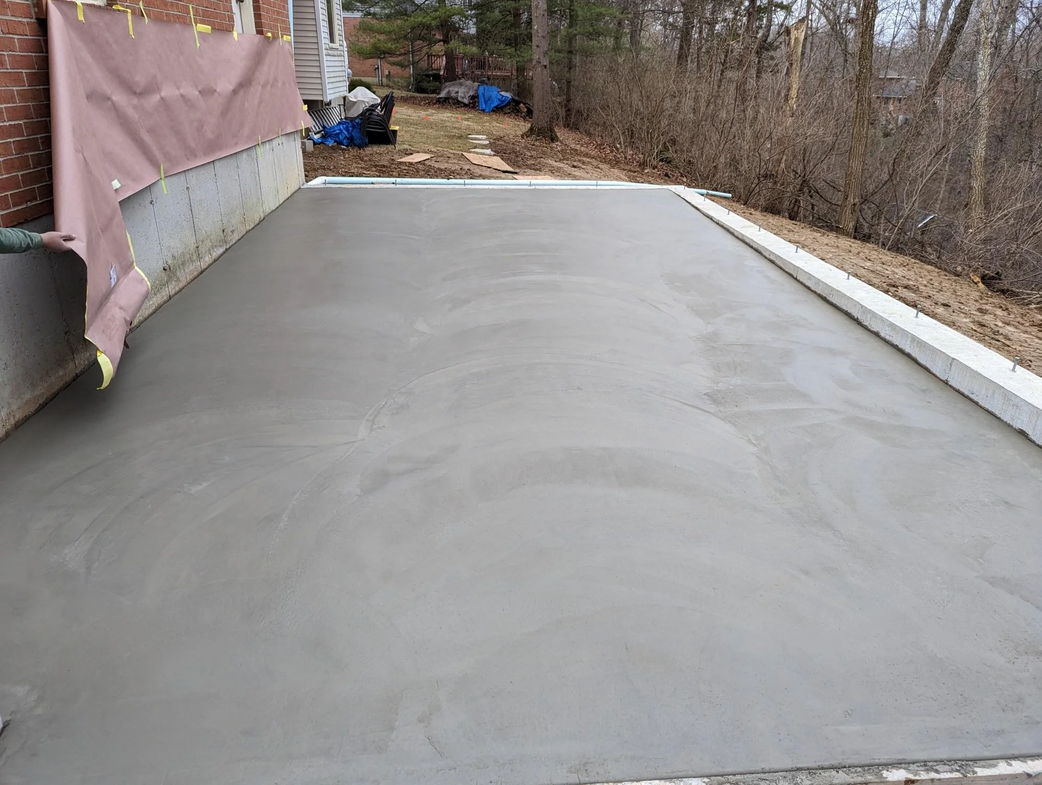 Photo of a freshly poured concrete patio or driveway area next to a house, with a curb, trees, and a backyard with some outdoor furniture and construction materials in the background.