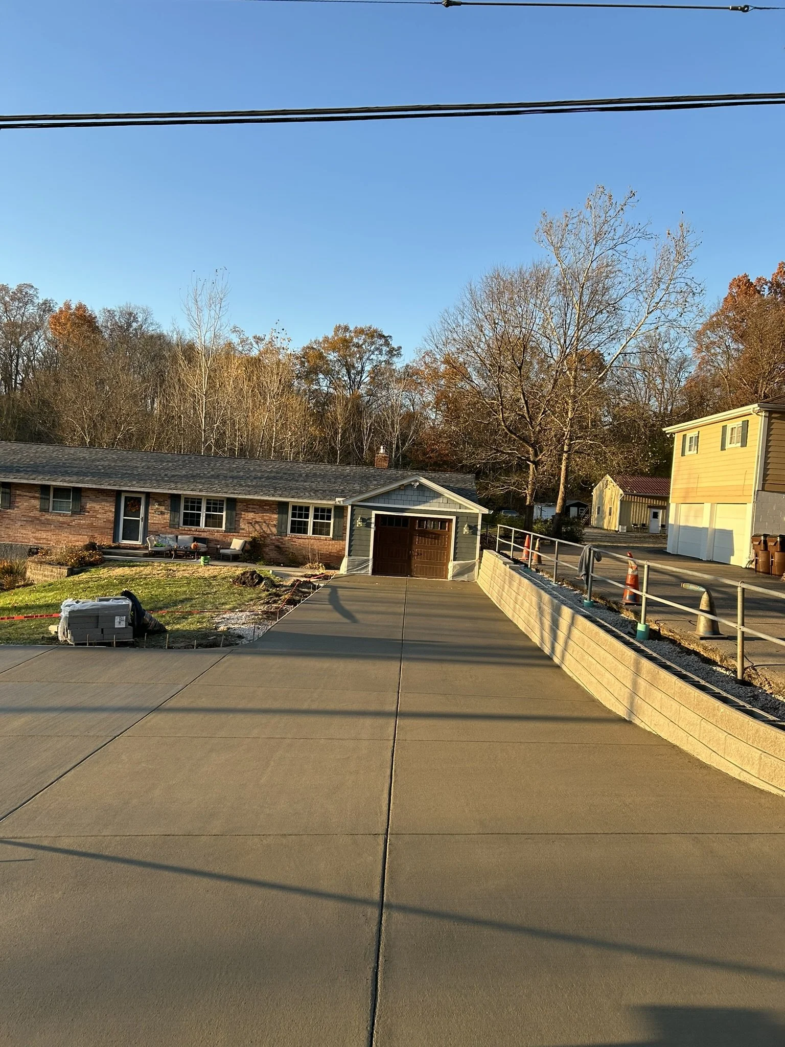 A newly paved concrete driveway leading to a garage attached to a brick house, with a lawn and some outdoor furniture on the side. There are trees with autumn foliage in the background and a clear blue sky overhead.