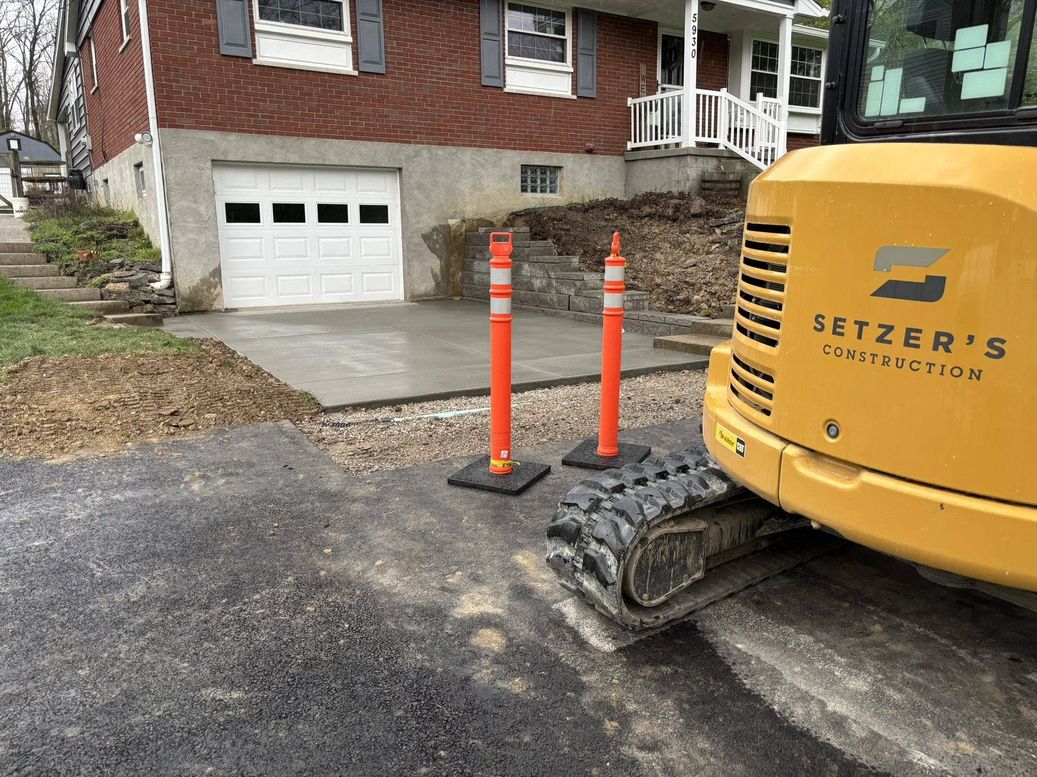 Construction site in front of a brick house with a white garage door, orange traffic cones, a yellow construction vehicle, and a newly paved concrete driveway.