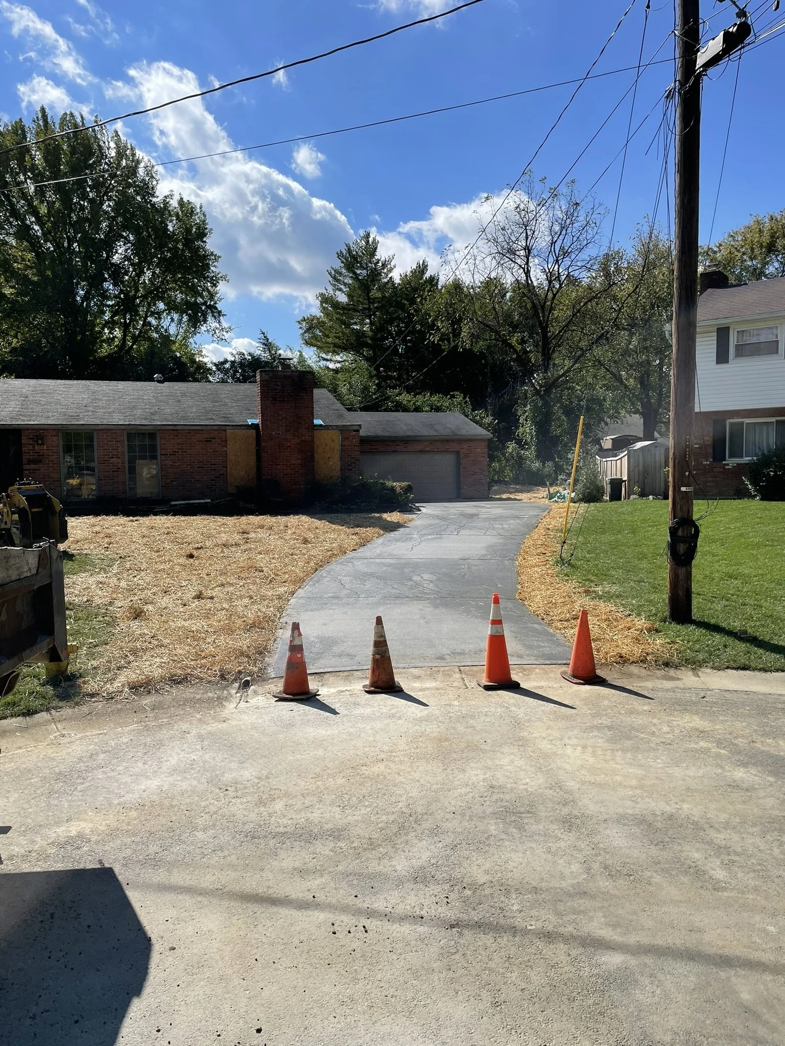 A residential driveway under construction with orange traffic cones blocking the entrance. The driveway is newly paved and leads to a brick house with a chimney. There are two houses on either side, and a utility pole with wires is visible on the rig