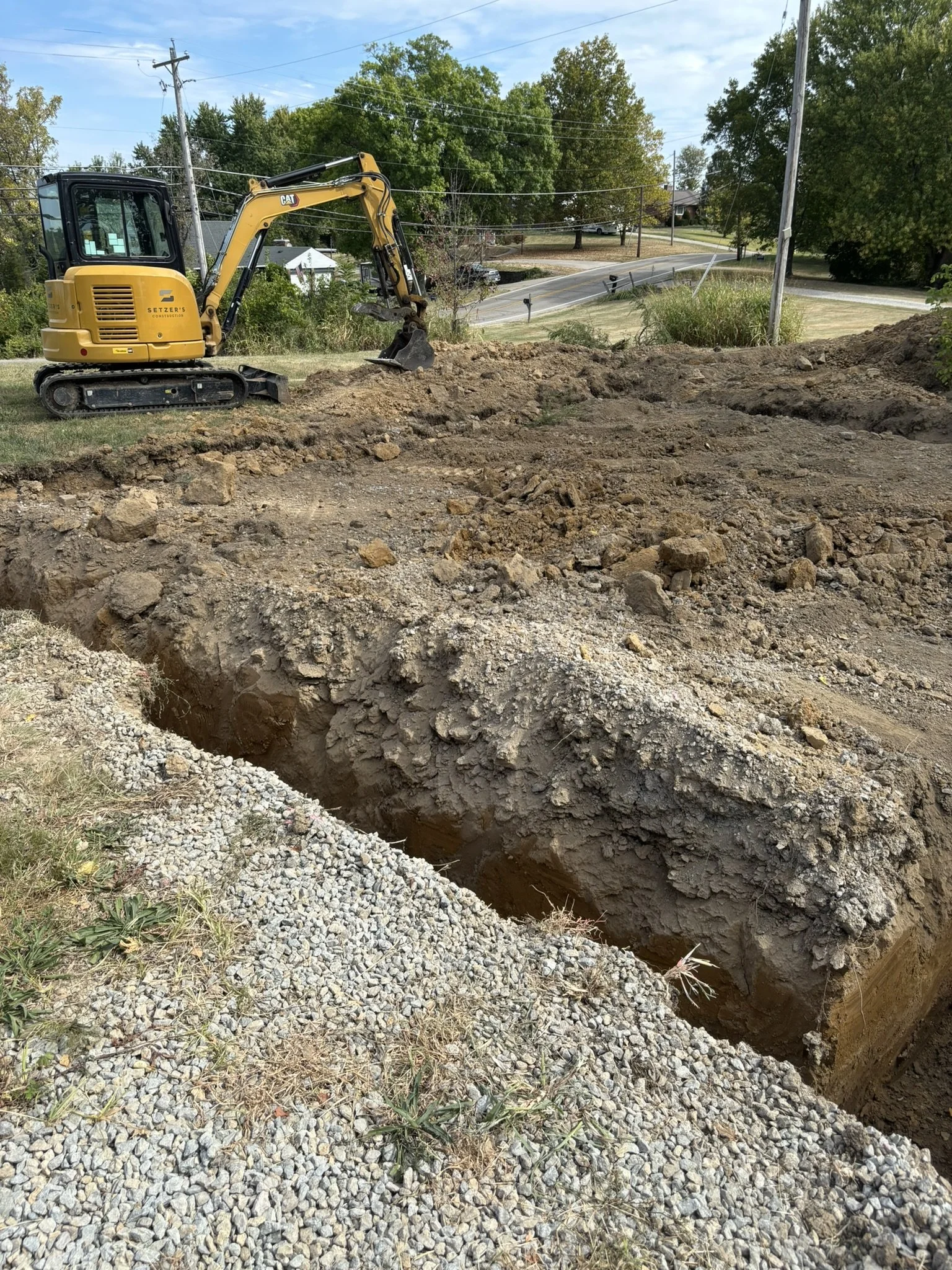 A yellow mini excavator digging a trench in a grassy area near a road, with trees and power poles in the background.