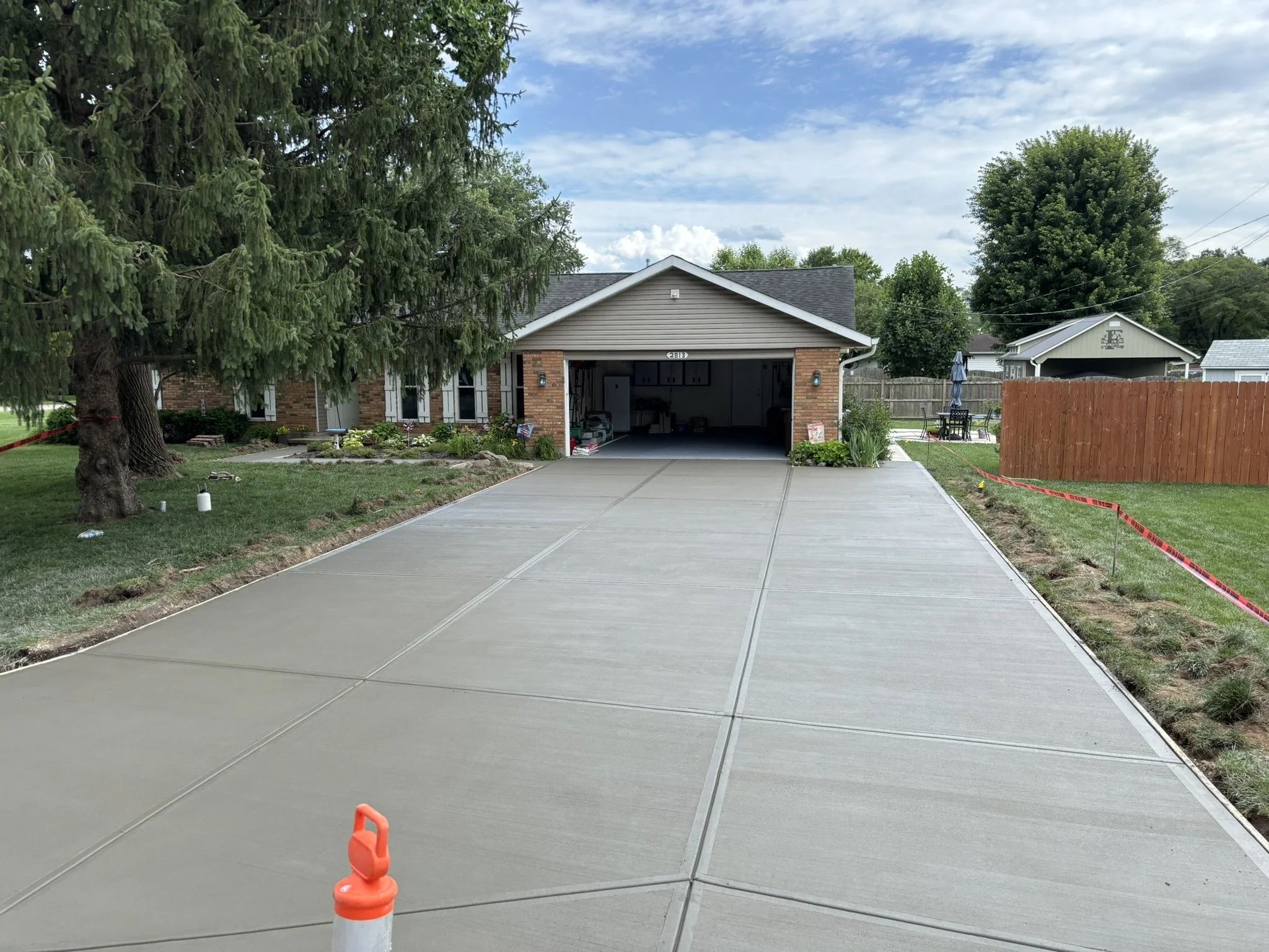 A newly poured concrete driveway leading up to a house with an open garage. The house has tan siding, brick accents, and a gabled roof. A large evergreen tree is on the left, and there is a backyard with a wooden fence, outdoor furniture, and a shed 