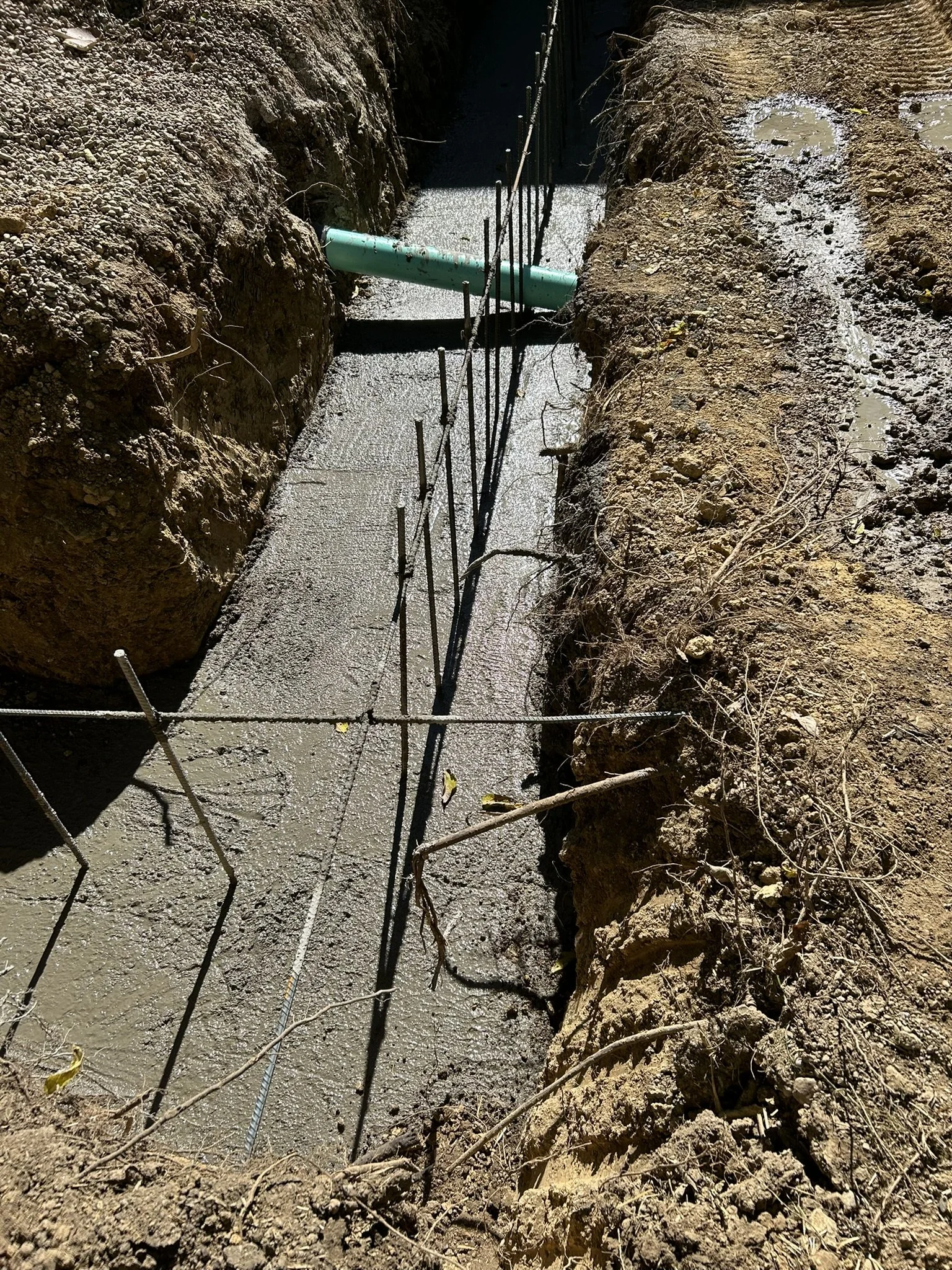 A construction site with a recently poured concrete foundation, a green pipe running through a trench, and steel rebar sticking out of the ground.