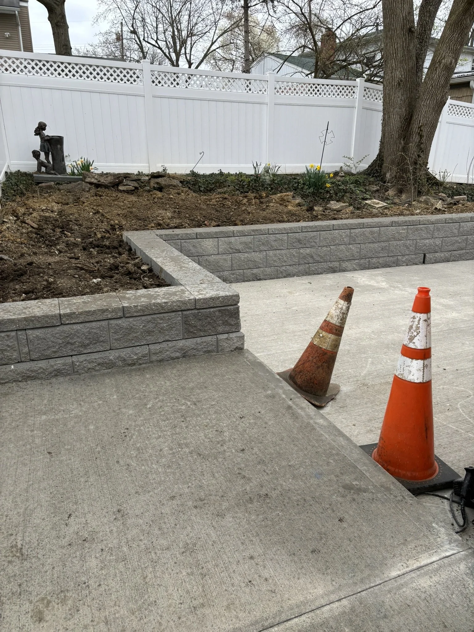 Construction zone with two orange traffic cones on concrete sidewalk, a gray brick wall, a dirt area, and a white privacy fence with trees and houses in the background.