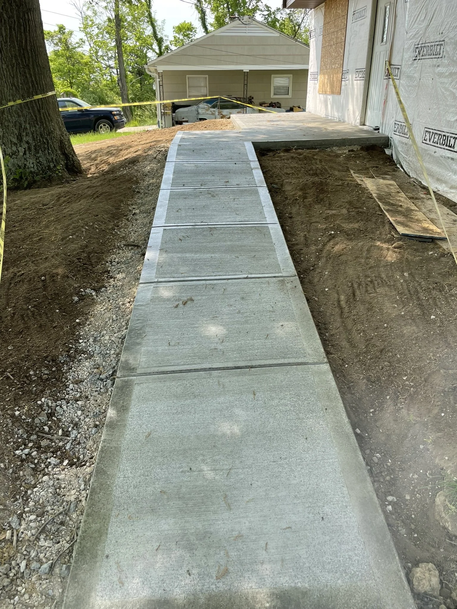 Concrete sidewalk under construction leading to a house, with soil on the sides and construction materials nearby.