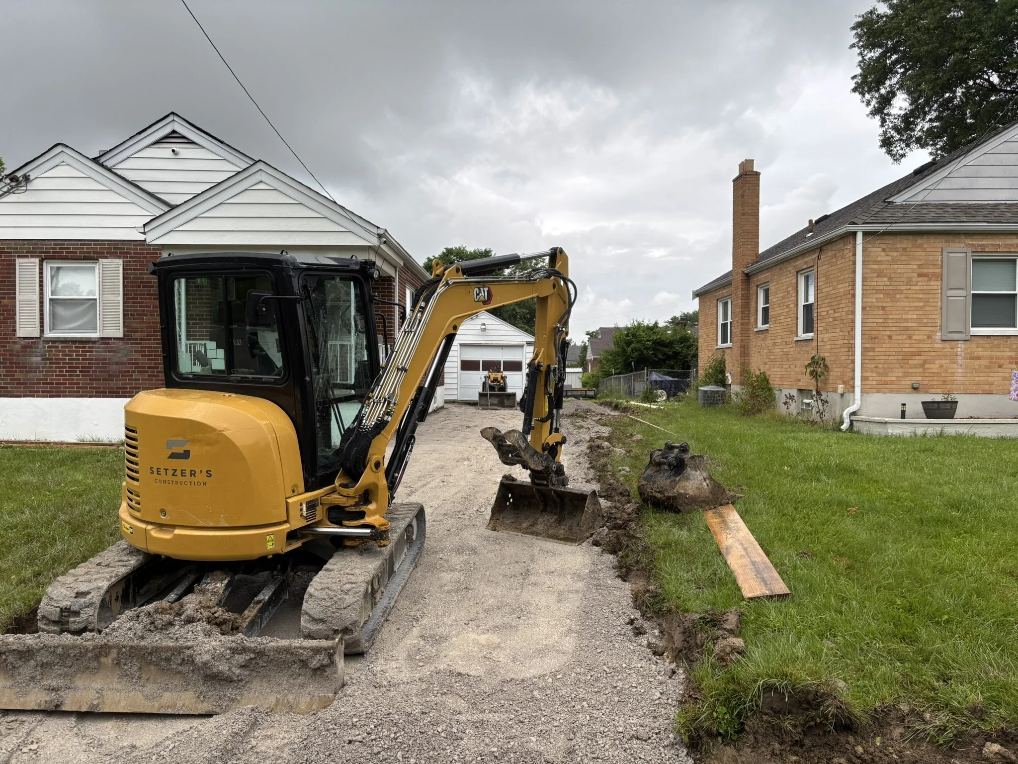 A yellow CAT mini excavator is digging a trench in a backyard, with two houses visible on either side and a cloudy sky above.