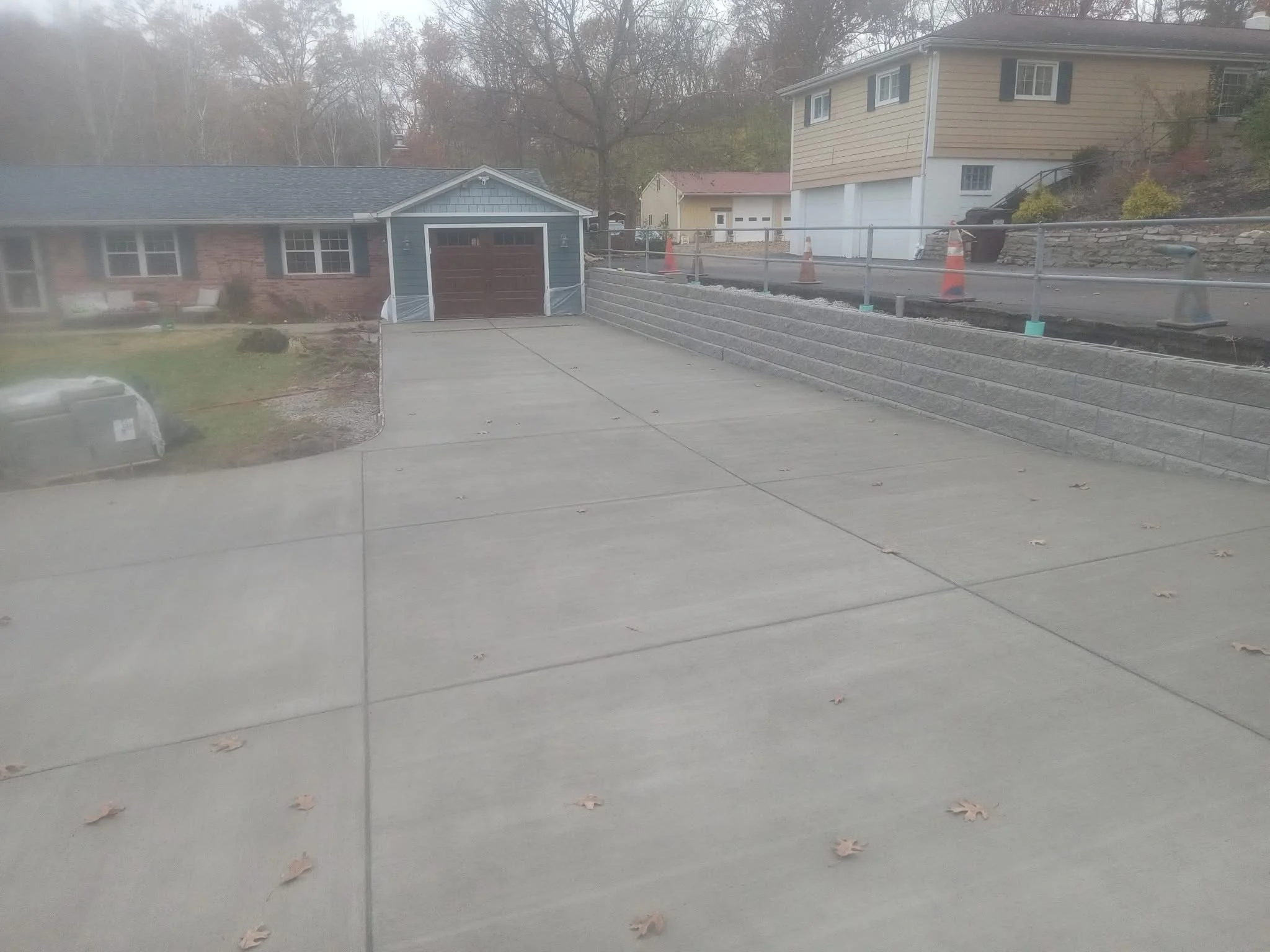 View of a driveway leading to a backyard with a blue house to the left and a beige house to the right, with trees and a cloudy sky in the background.