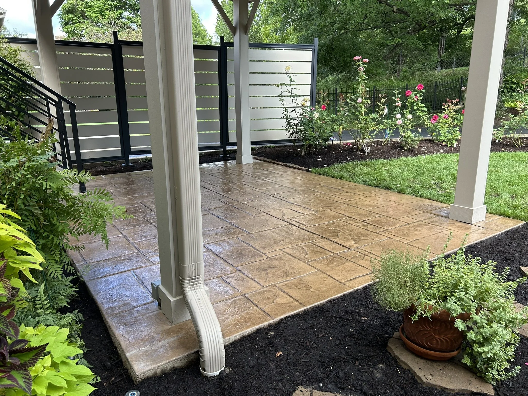 A backyard porch with brown stone tile flooring, white columns, a black and white privacy fence, rose bushes, potted plants, and greenery with trees in the background.