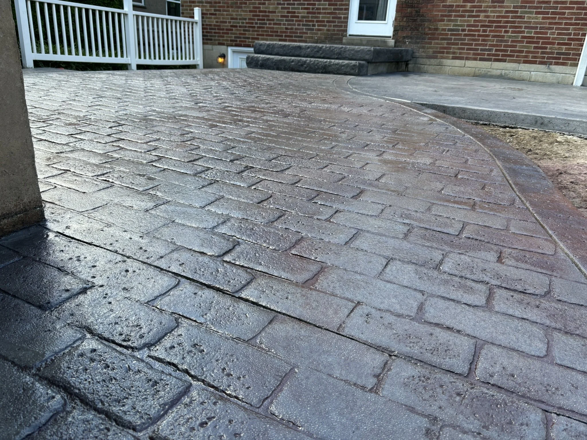 Close-up of a brick house entrance with stone stairs, a door, a white railing, and a paved walkway that appears to be wet.