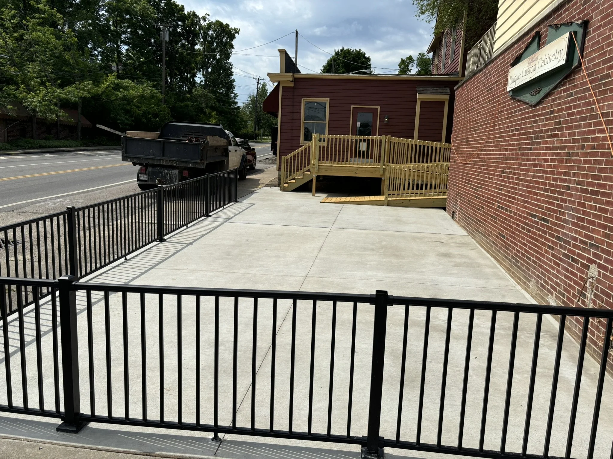 Empty sidewalk with black metal fence, brick building, and a small wooden ramp leading up to a door, parked truck on the street, trees and cloudy sky in the background.