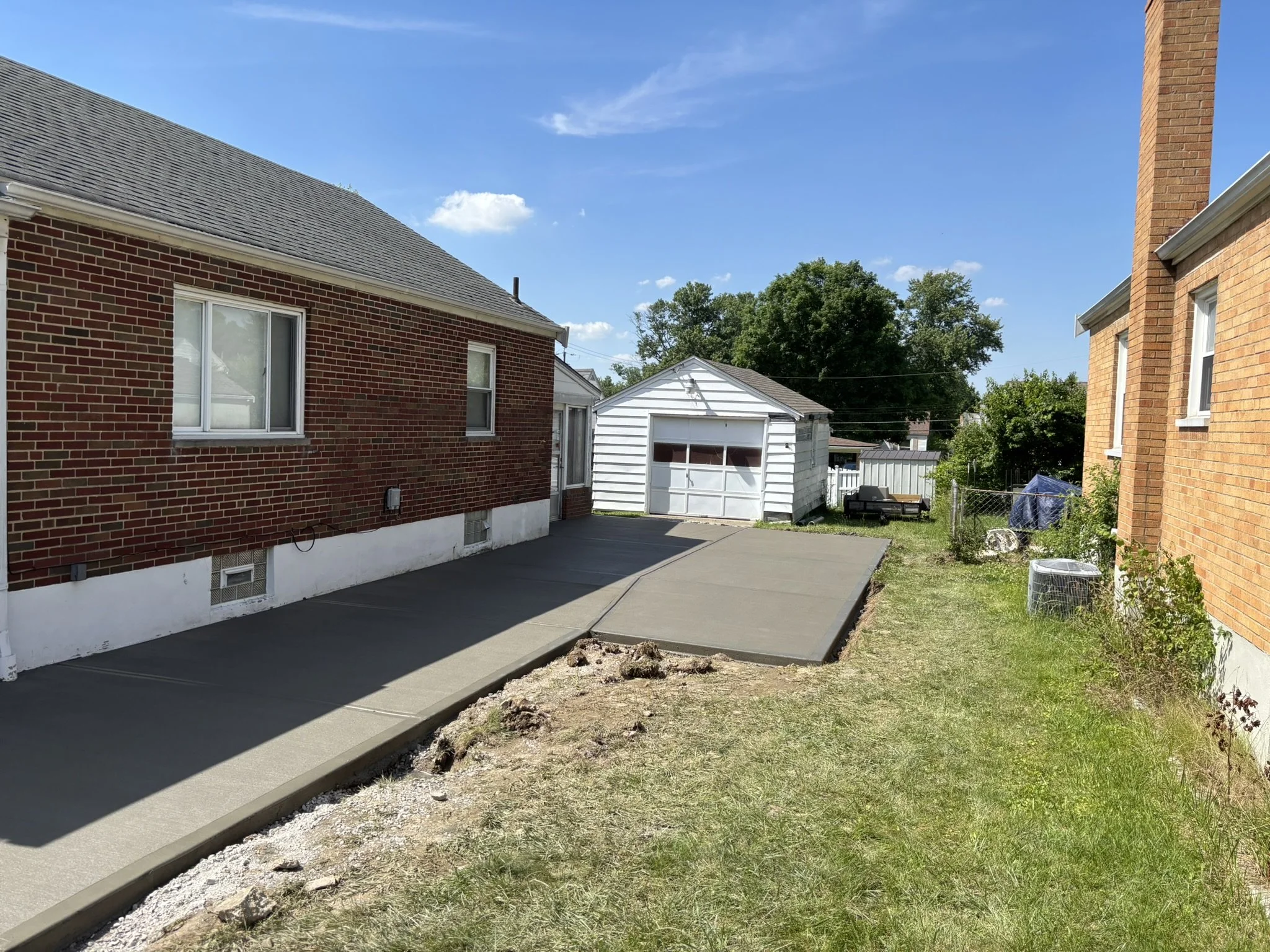 A backyard with a fresh concrete patio under construction next to a brick house, with a white shed, green trees, and a blue sky with some clouds.