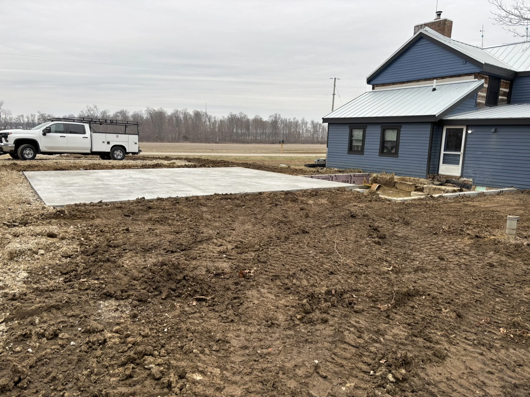 A construction site showing a blue house with a metal roof, unfinished foundation, and a large white utility truck parked nearby, with a field and trees in the background on a cloudy day.