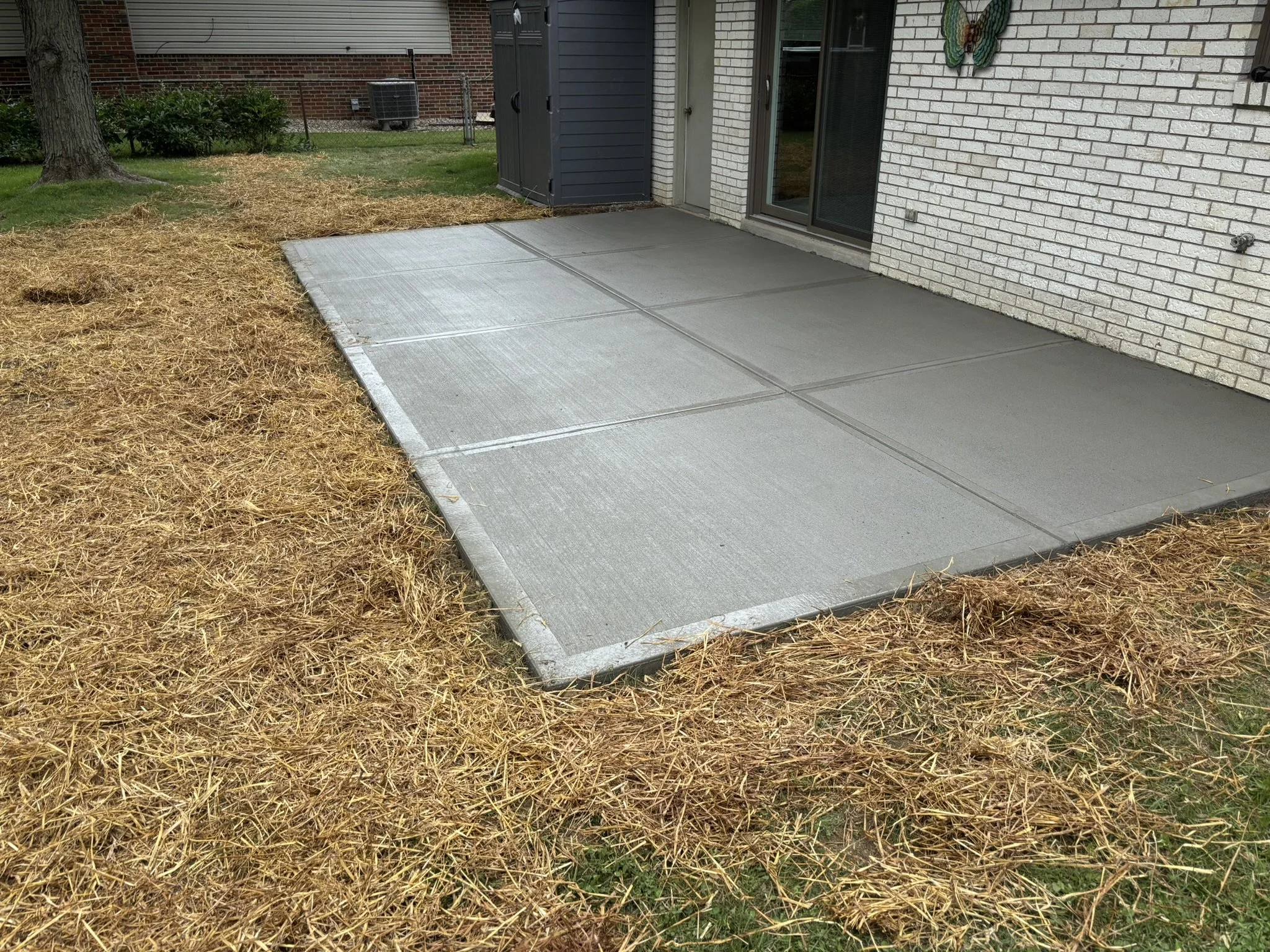 Concrete patio area beside a brick house with a white textured wall, a black shed, a sliding glass door, and a decorative butterfly on the wall. Surrounding the patio is brown mulch and green grass, with a tree trunk and shrubbery visible.