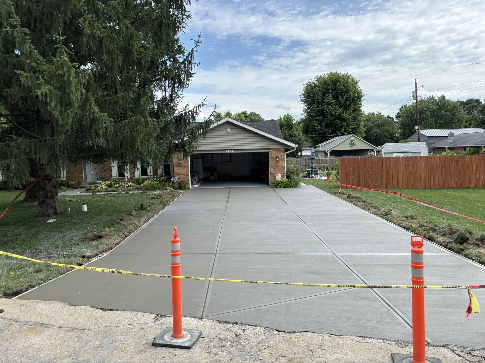 Freshly poured concrete driveway section with orange traffic cones and caution tape, surrounding a residential house with a garage and outdoor patio area, under a partly cloudy sky.