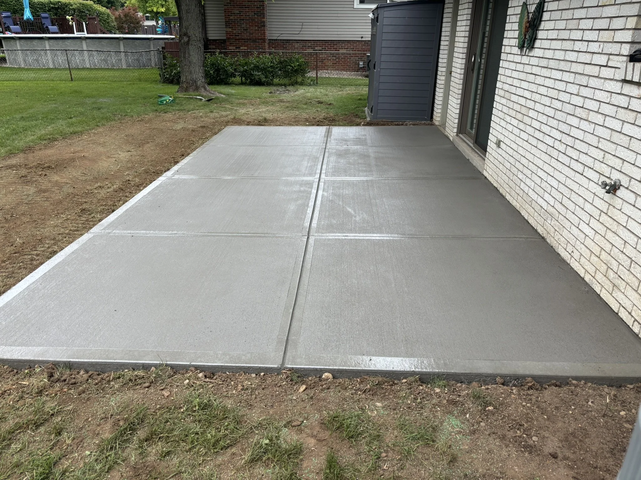 Patch of soil next to a newly poured concrete patio in a backyard, with a tree and grass in the background.