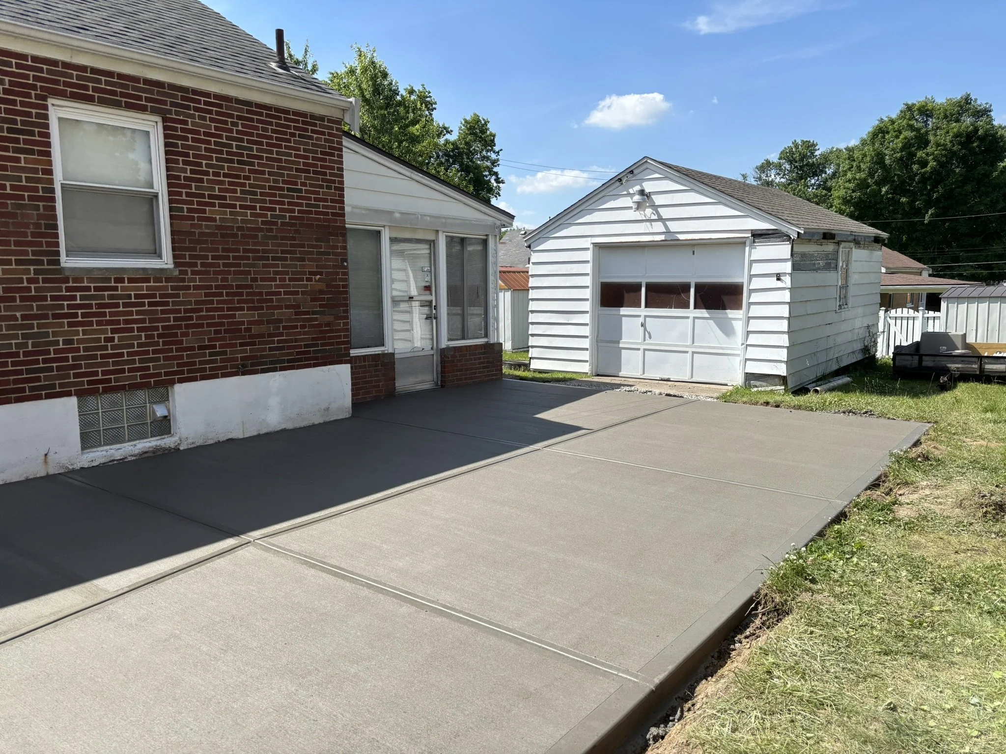 A backyard scene with a red brick house on the left and a white garage on the right. The sky is blue with some clouds, and there are trees in the background. There is a concrete patio in the foreground and a grassy area with some soil along the edge.