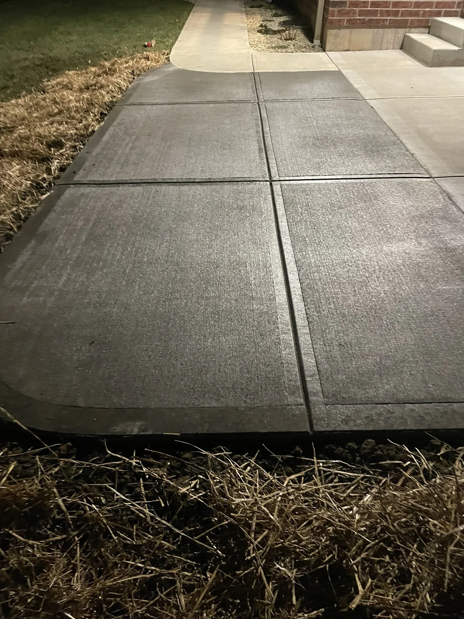 Nighttime photo of a concrete sidewalk near a house with stairs, bordered by a patch of grass and dried plants.