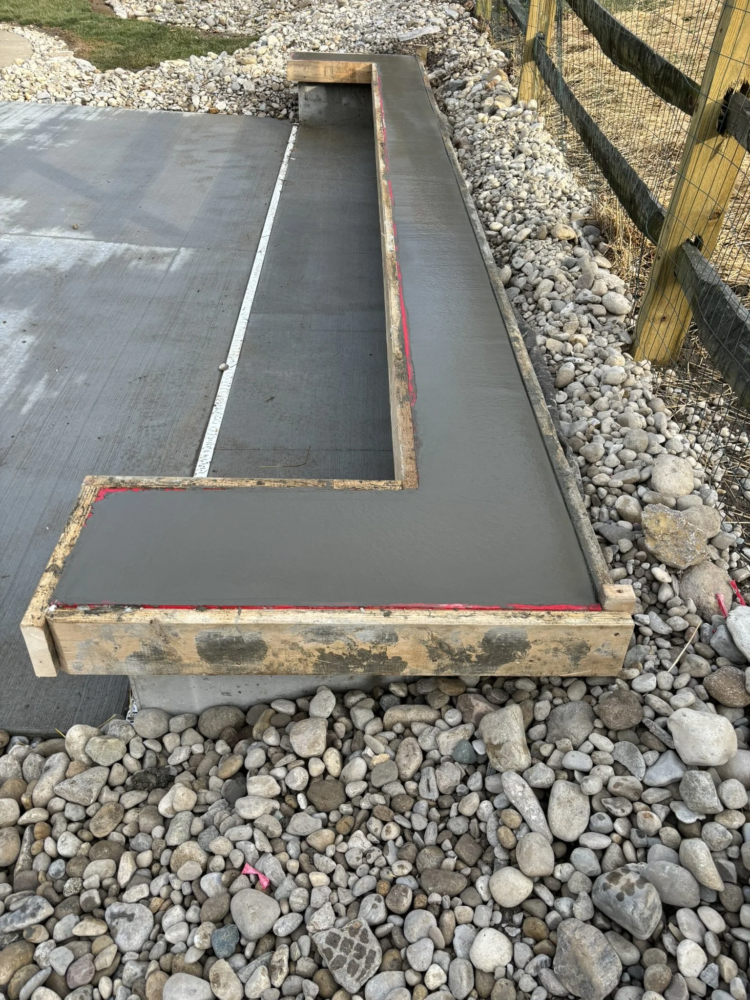 A concrete skate ramp with a coping edge, surrounded by small rocks and gravel, next to a wooden and wire fence.