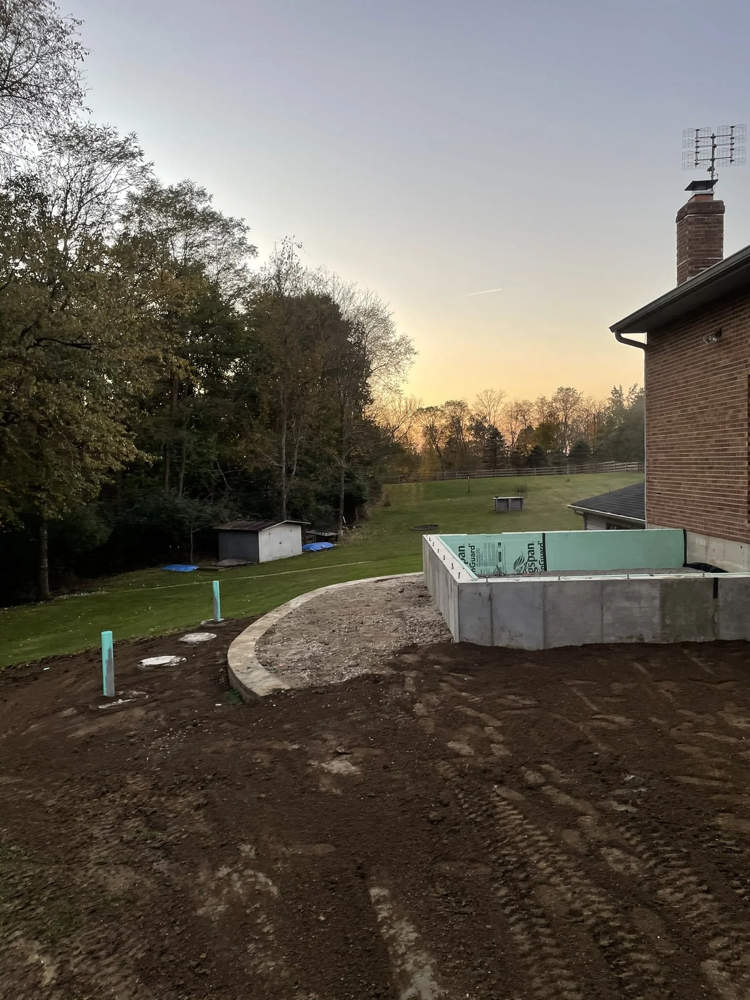 A backyard under construction with a dug-up dirt area, a small concrete wall, a house with a brick chimney, a green fence, trees in the background, and the sky at sunset.