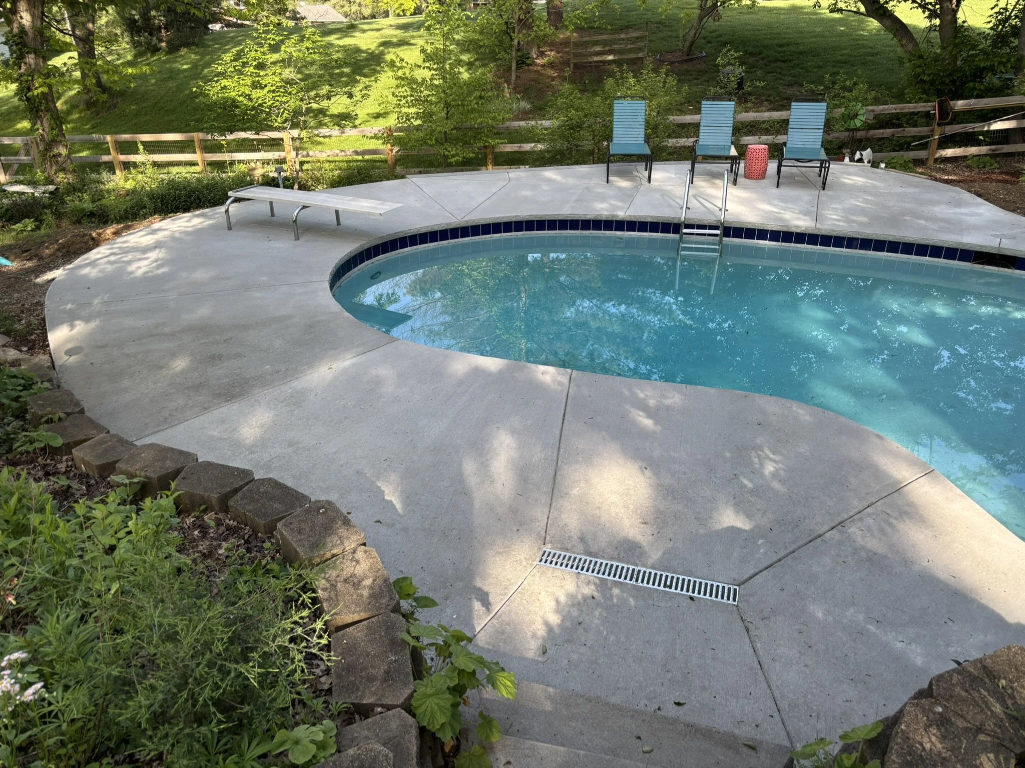 Backyard patio with concrete surface, pool with blue water, steps with metal handrails, and three blue lounge chairs next to a pink side table. Green grass, trees, and a wooden fence in the background.