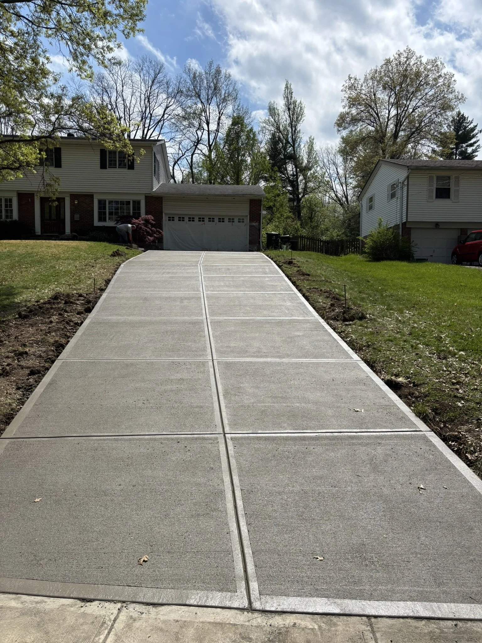 Freshly poured concrete driveway leading up to two houses, with a small flower bed on the left, and trees in the background under a partly cloudy sky.