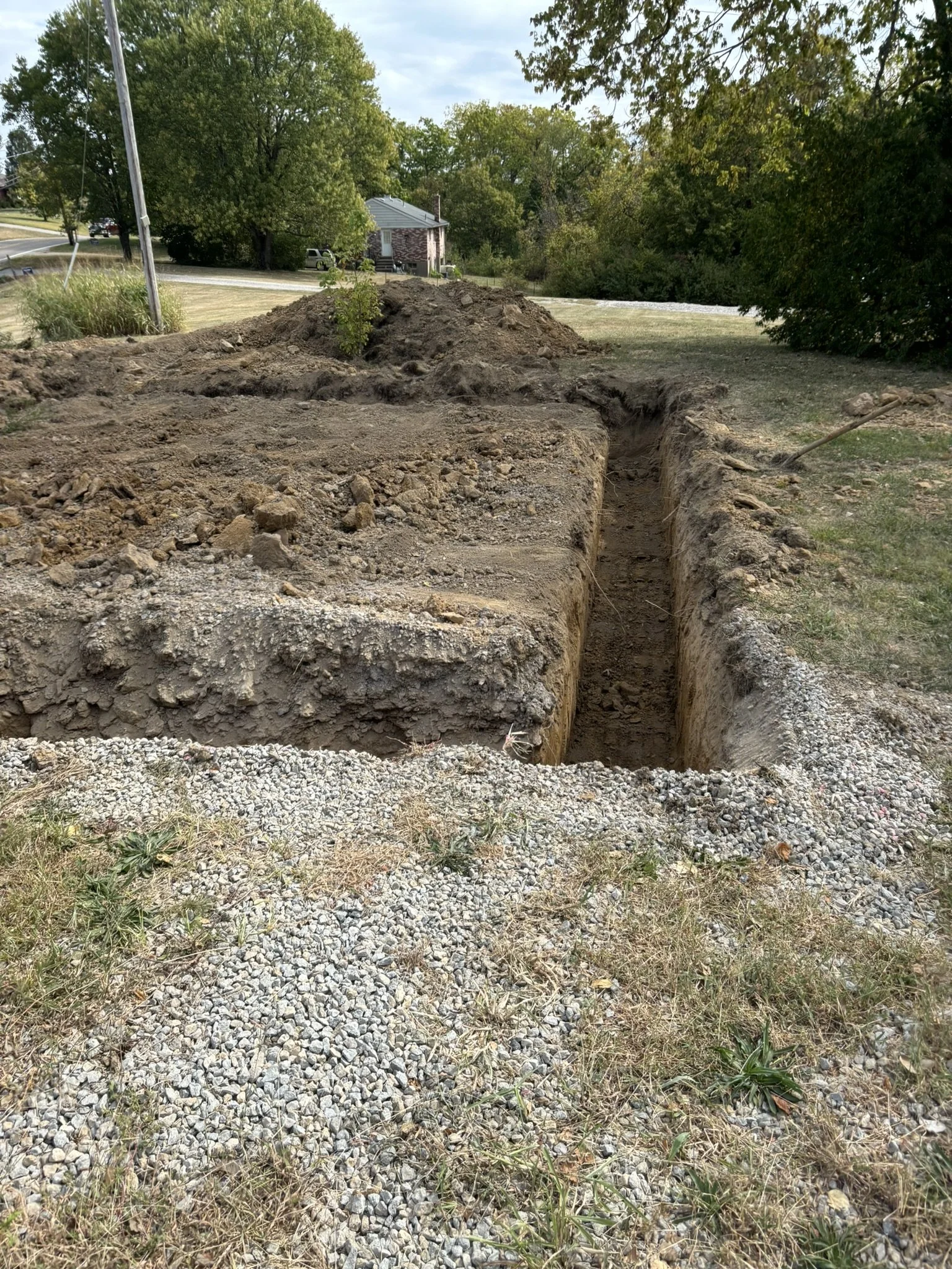 A dug trench in the ground with dirt mounds on either side, surrounded by grass and gravel, with trees and houses in the background.