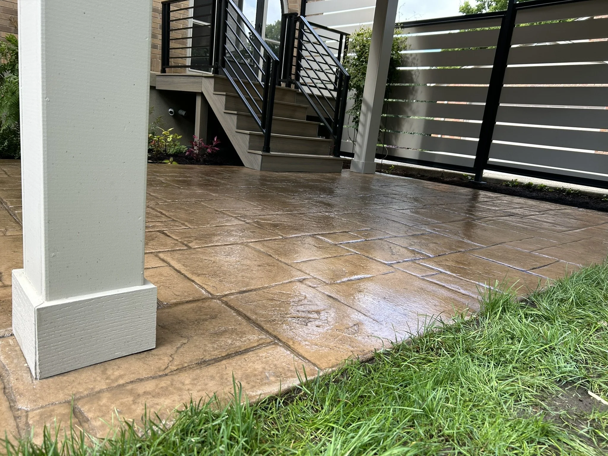 A porch area with wet, brown stamped concrete flooring, a staircase with dark metal railings leading up to a door, and a beige painted pillar in the foreground. There are small plants and grass along the edge of the porch.