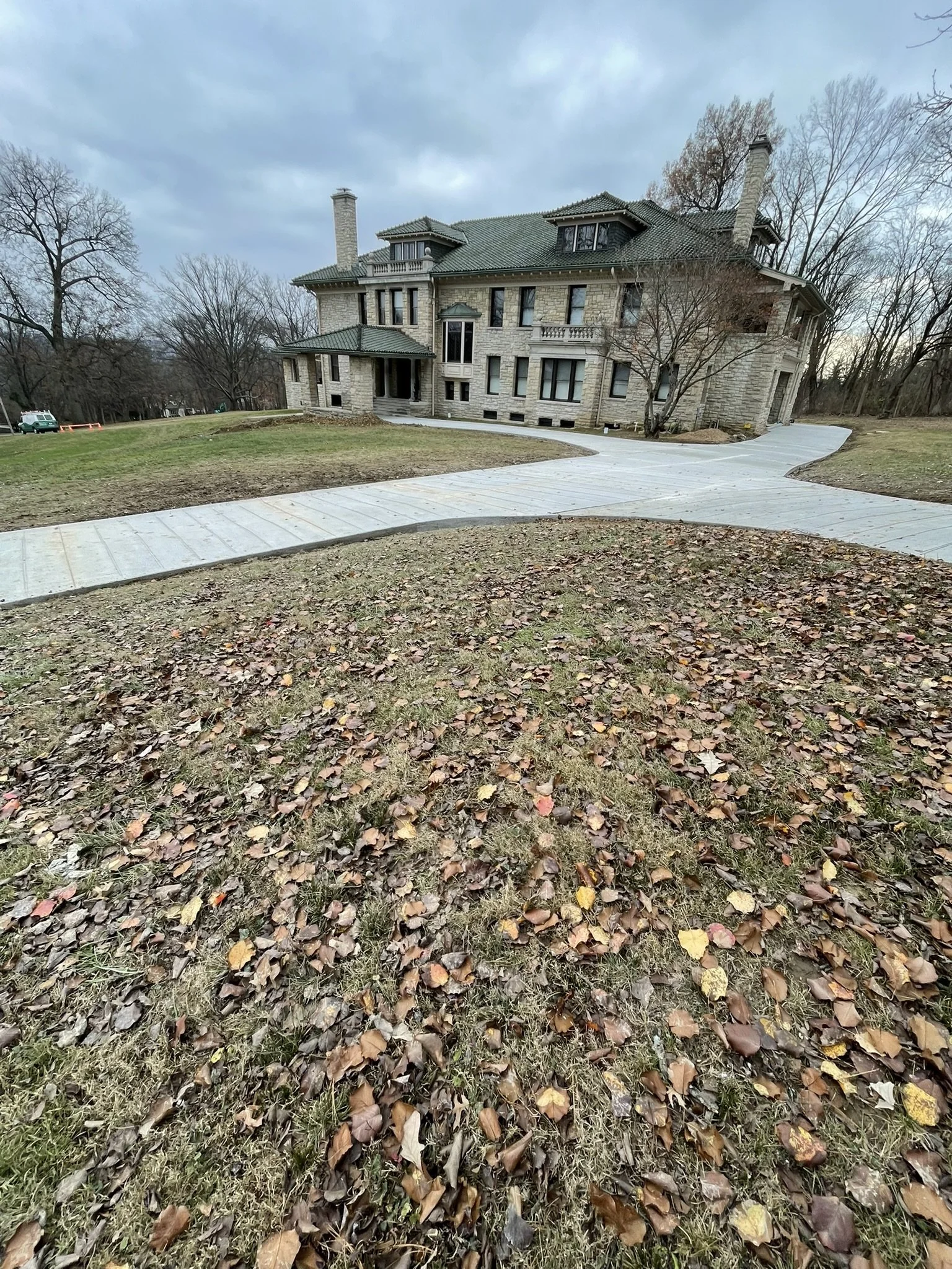 A large house with stone exterior walls, multiple chimneys, and a green tiled roof, surrounded by leafless trees and a lawn with fallen leaves, under a cloudy sky.