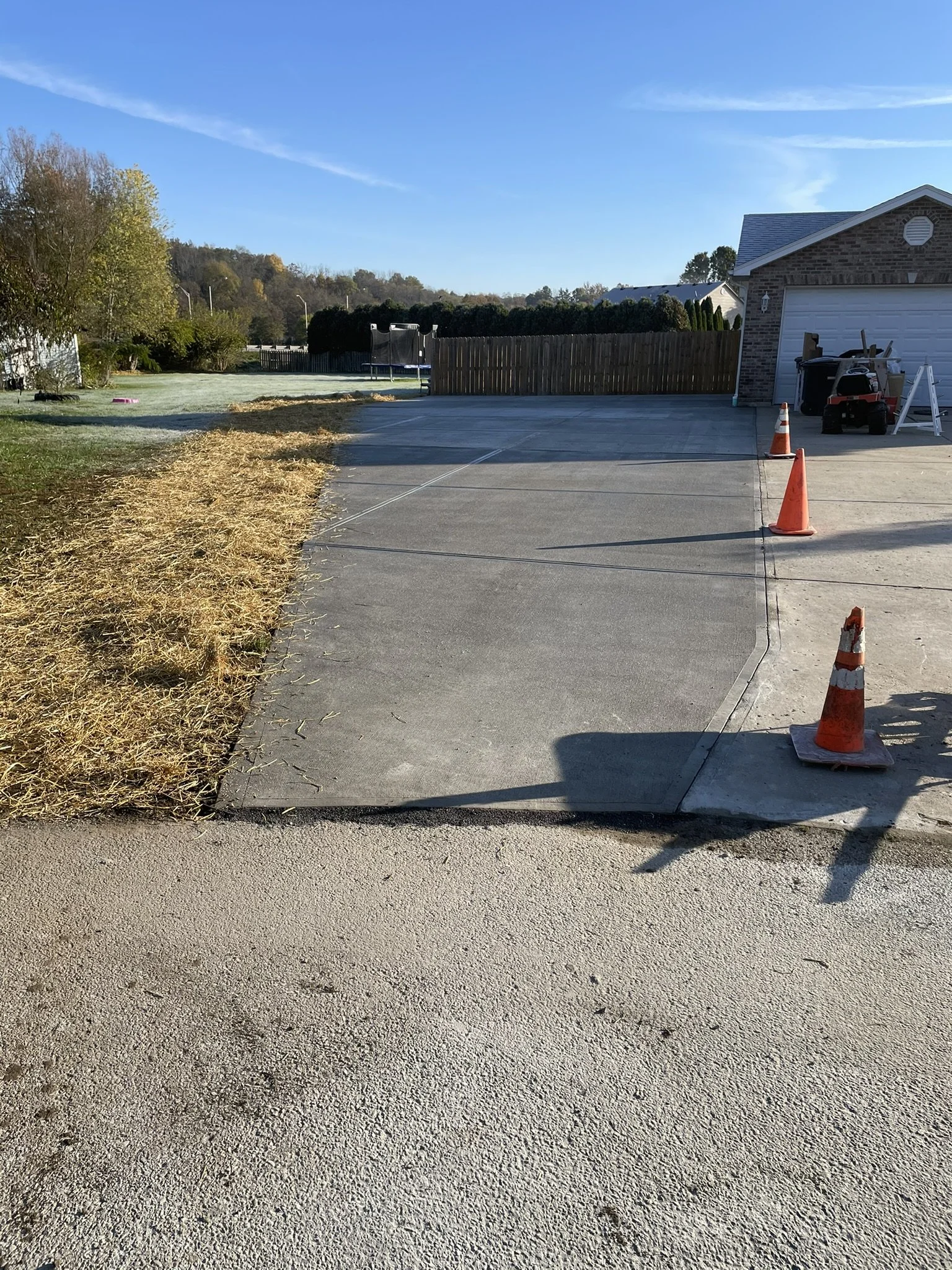 A driveway with orange construction cones, a trash bin, and a small vehicle parked in front of a brick house with a garage door. To the left, there is a yard with grass and some straw; to the right, a wooden fence and some trees in the background.