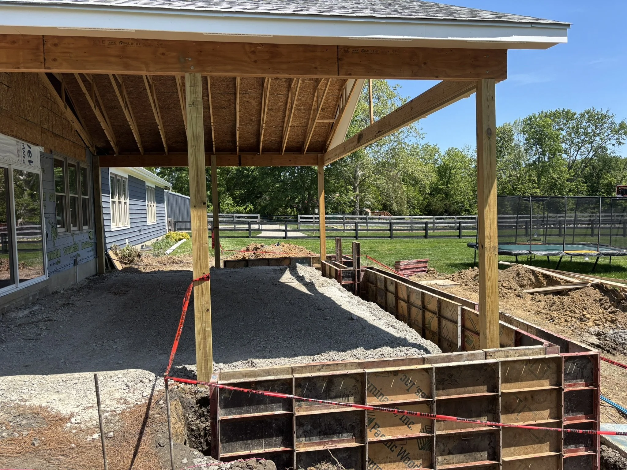 Under construction backyard patio area with wooden support beams, concrete foundation, and a partially built roofing structure. There are trees, a blue house, and a trampoline in the background.