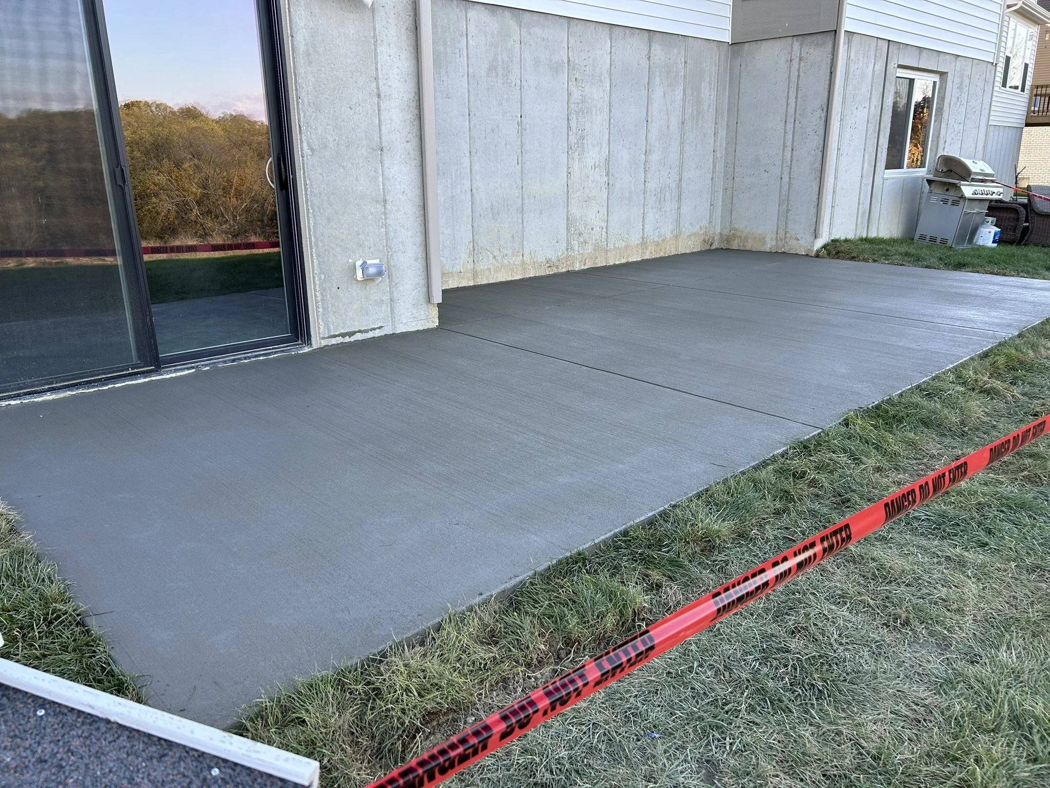 Outdoor concrete patio area with sliding glass door, a wall-mounted outdoor electrical outlet, and a barbecue grill. There is caution tape on the grass nearby.
