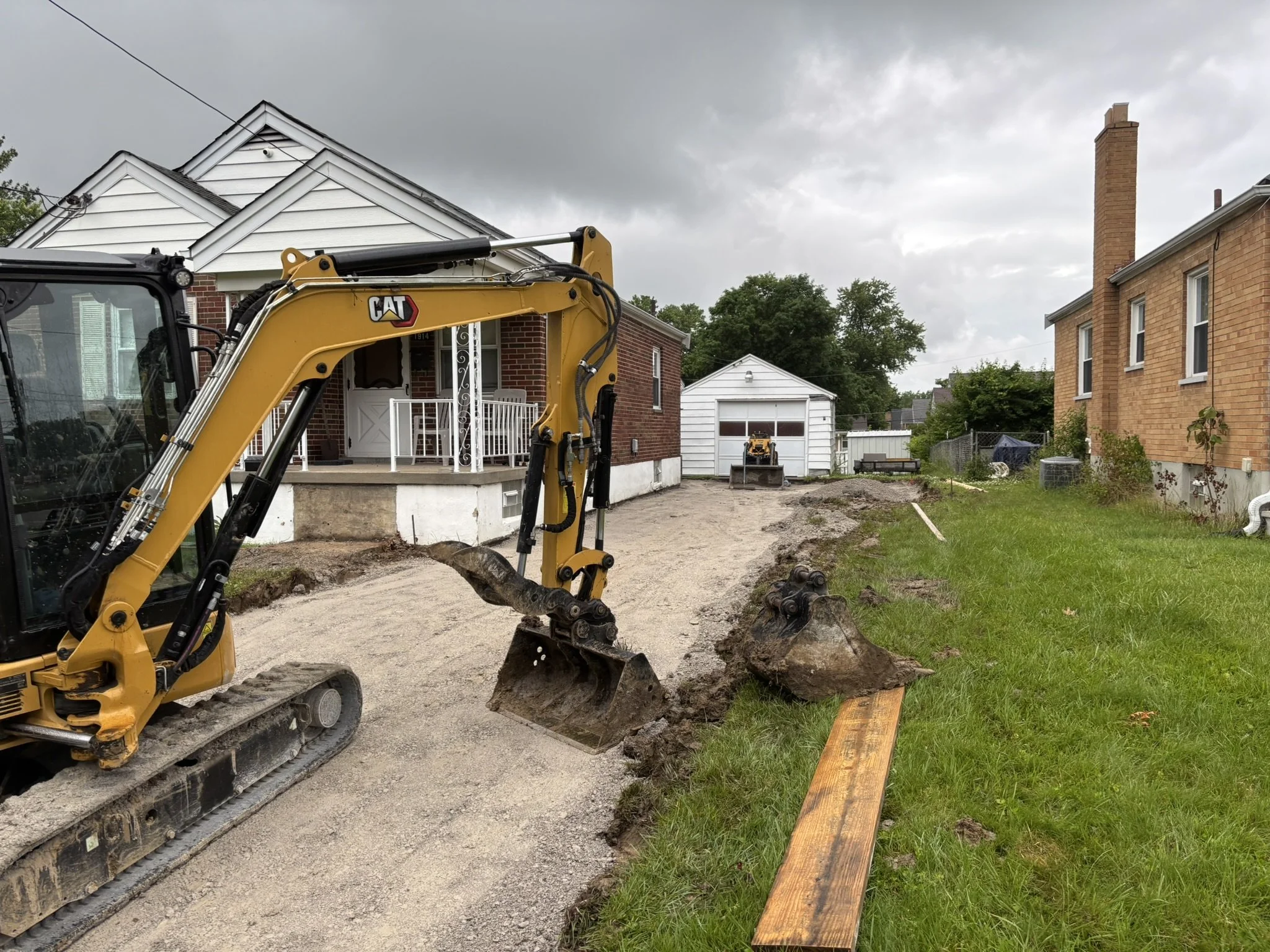 A yellow Caterpillar mini excavator digging a trench in a backyard with houses and a cloudy sky in the background.