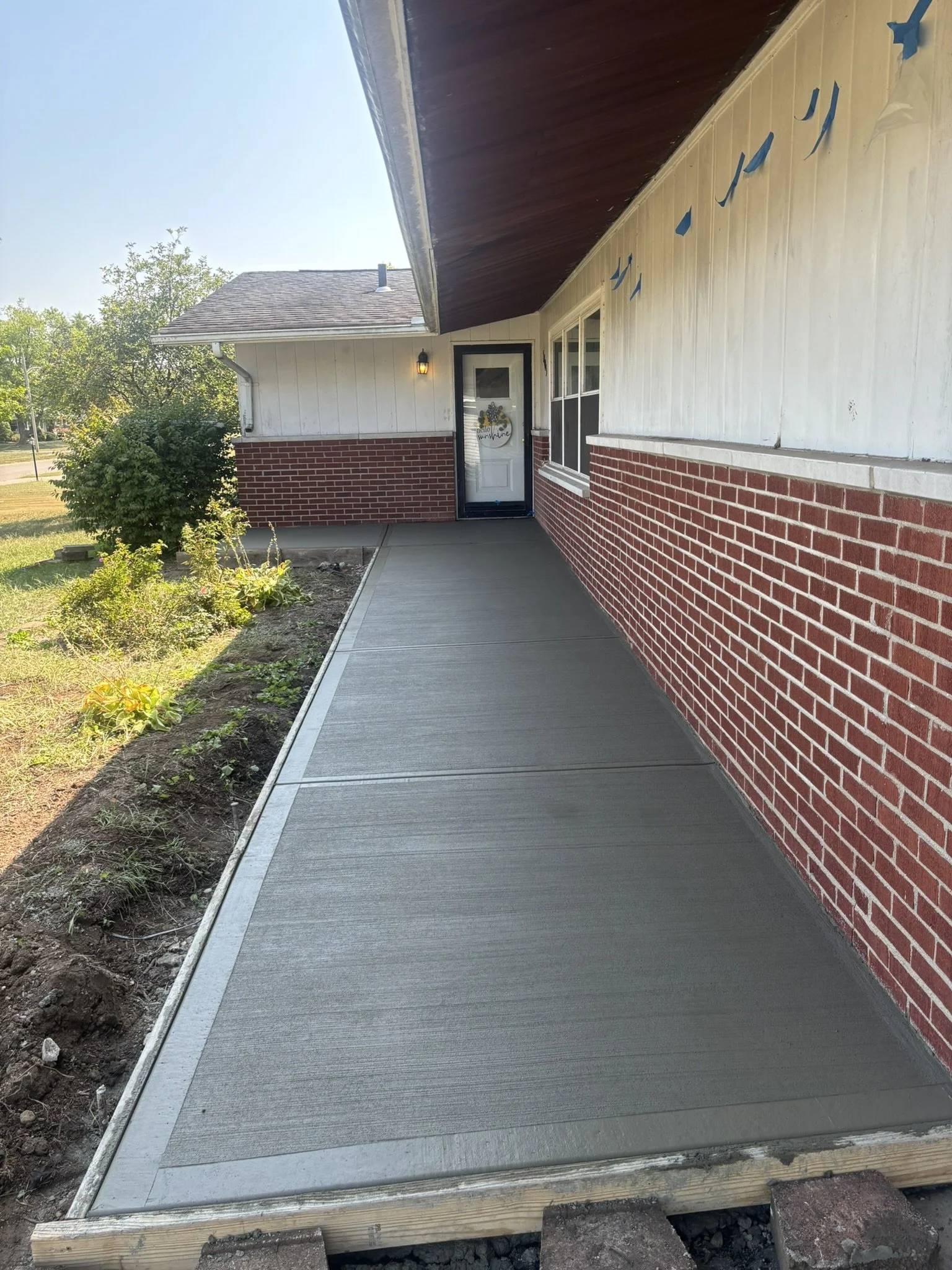 Concrete sidewalk in front of a house with a brick wall exterior and a white door, plants, and trees on the left, daytime with clear sky.