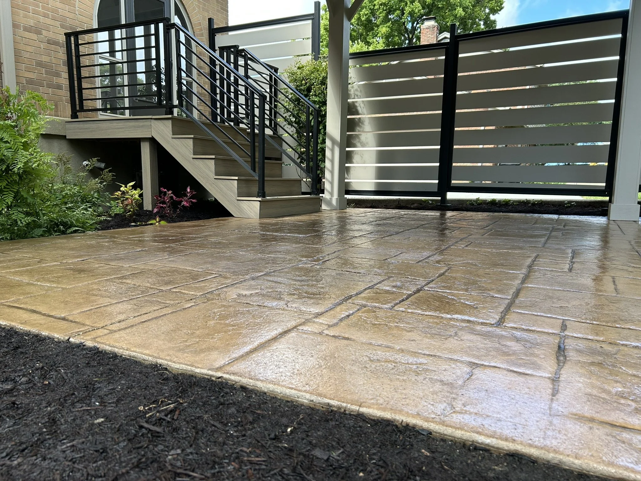 A backyard patio featuring stained concrete paving, a wooden staircase with a black metal railing leading up to a house, and a grey privacy fence with horizontal slats. There are plants along the side of the stairs and a partly cloudy sky overhead.