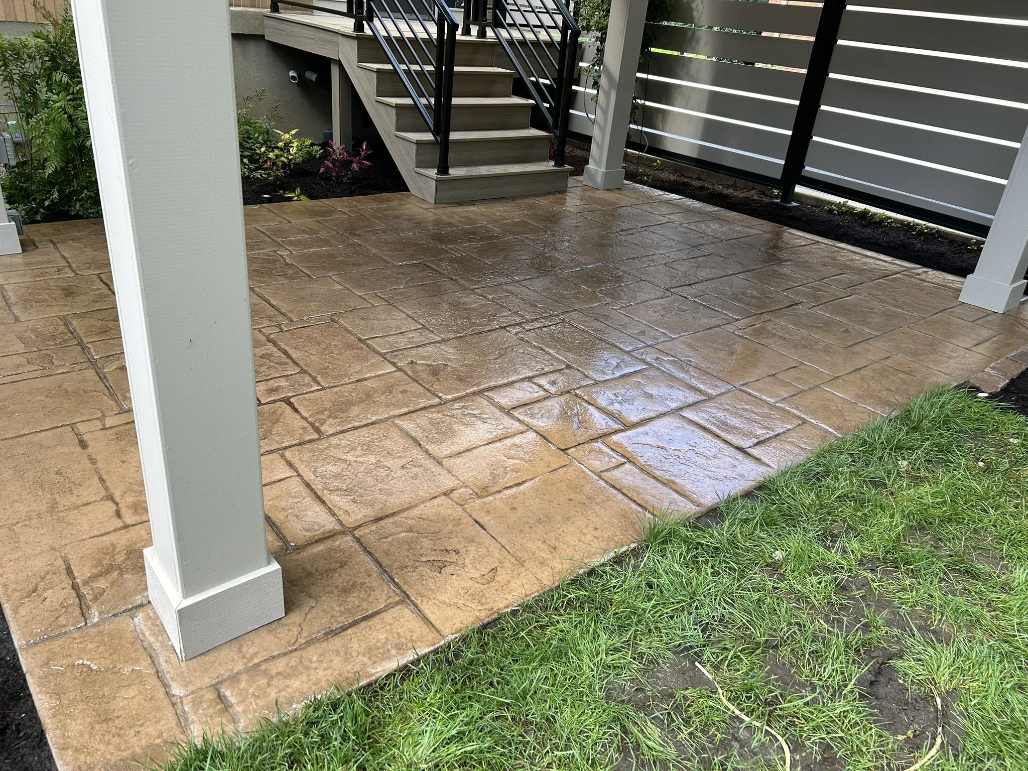 Rain-soaked brown stamped concrete patio with a grassy edge, metal staircase with black railings leading to a deck, and a white fence with horizontal slats in the background.